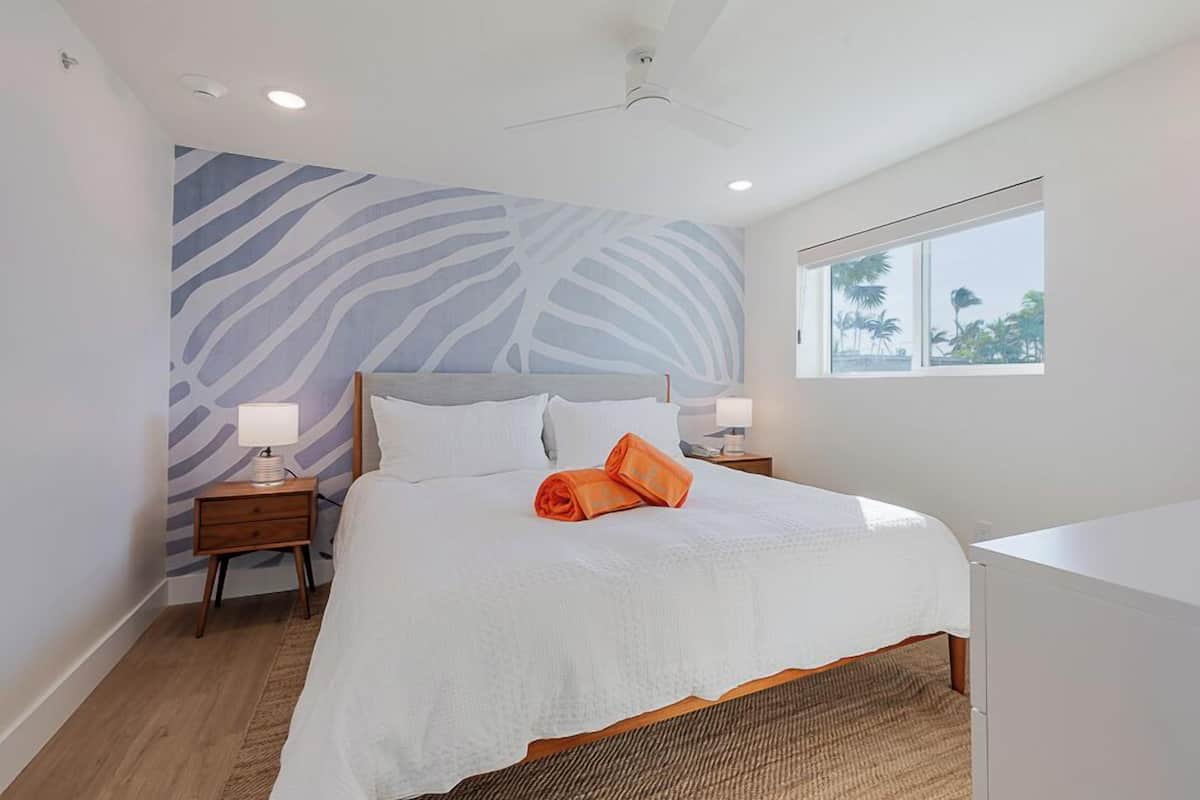 Ein Schlafzimmer mit einem großen Bett, zwei Nachttischen, einem Deckenventilator, einem Fenster mit Blick auf Palmen und einer Wand mit Zebra-Print-Design.