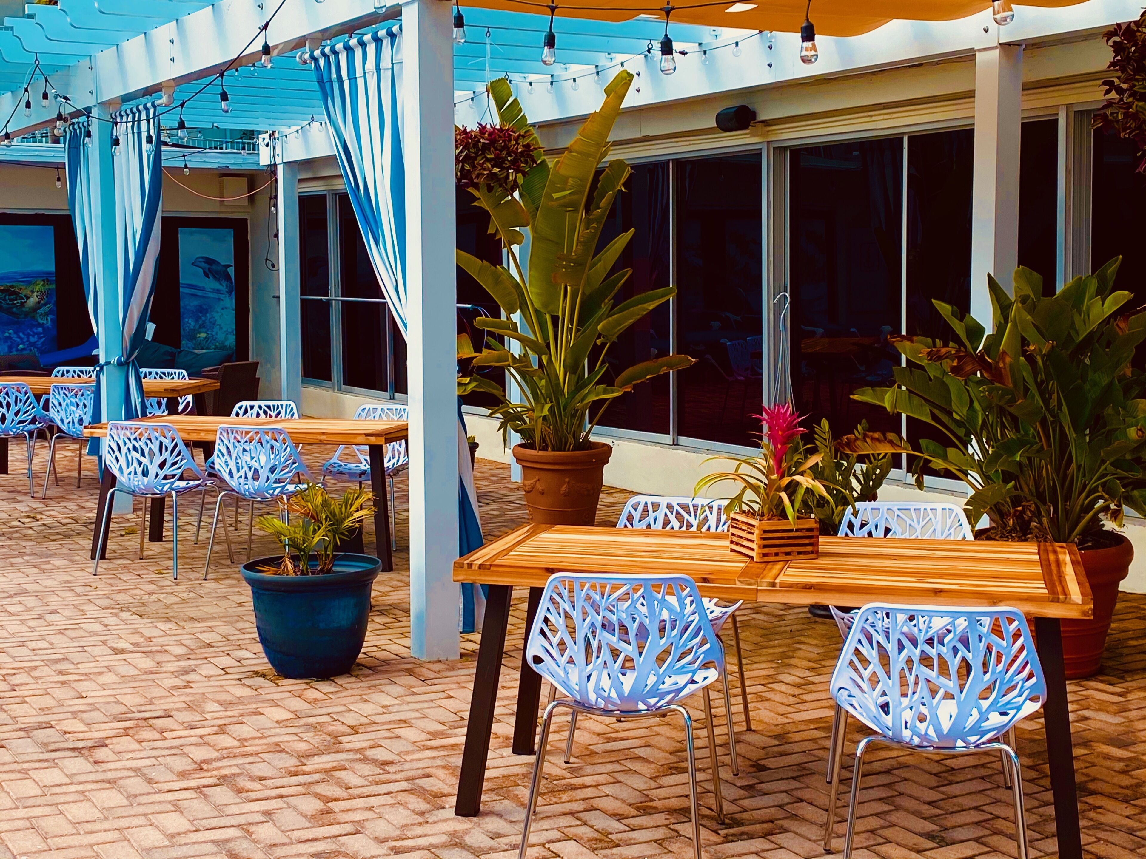 Uma área de pátio externa com mesas de madeira e cadeiras azuis, plantas em vasos e um espaço de estar coberto com cortinas azuis.