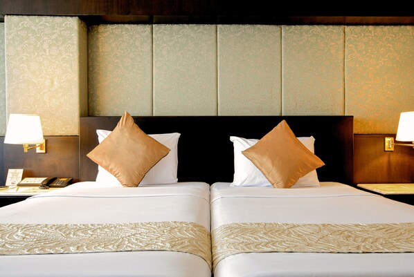 A hotel room with two beds, each with a headboard, white linens, and a decorative bed runner. There are two bedside lamps and a telephone on the left side of the bed.