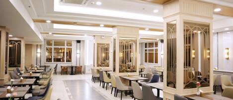 Een ruime hotel eetzaal met meerdere tafels en stoelen, grote ramen en decoratieve verlichting.