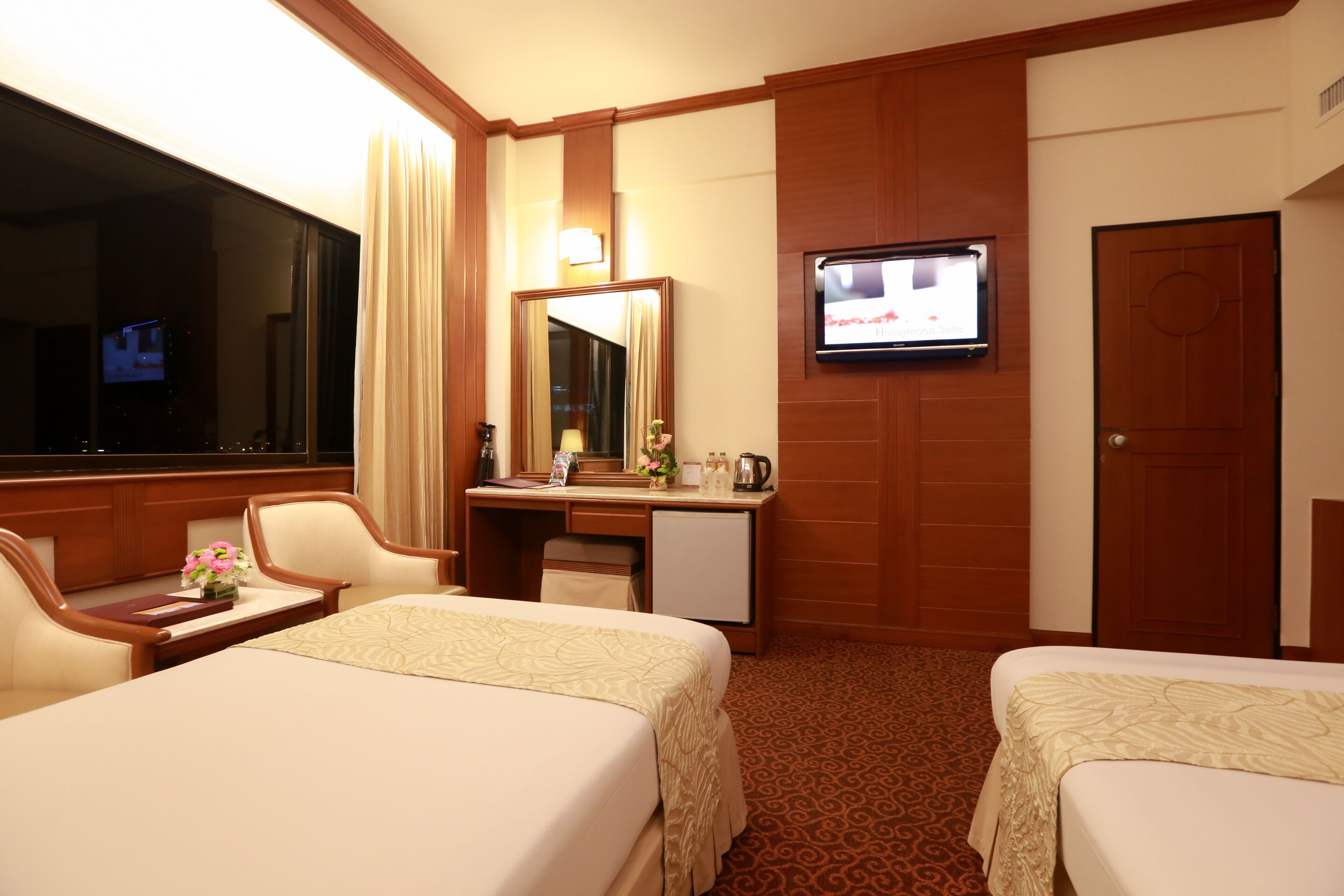 A hotel room with two beds, a desk with a mirror, a television mounted on the wall, and a door.