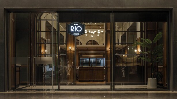Ein moderner Hauseingang mit Glastüren, einem Schild mit der Aufschrift "RIO Brazilian Fine Grill" und einer sichtbaren Kandelaber-Leuchte im Inneren.