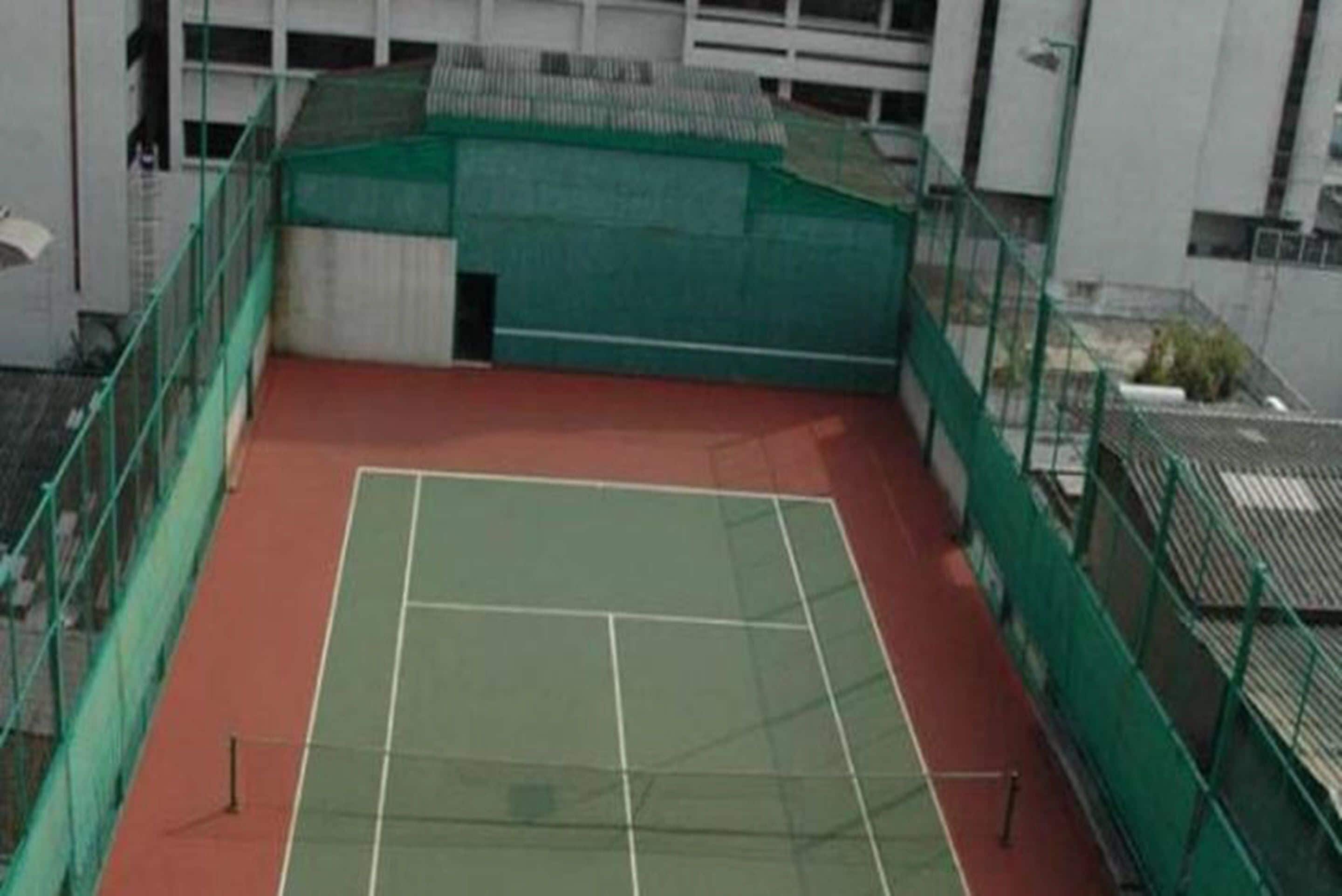 Ein Tennisplatz im Freien mit grünen und roten Belägen, umgeben von grünem Zaun, neben einem Gebäude mit weißer Fassade.