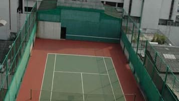 An outdoor tennis court with green and red surfaces, surrounded by green fencing, and adjacent to a building with a white facade.