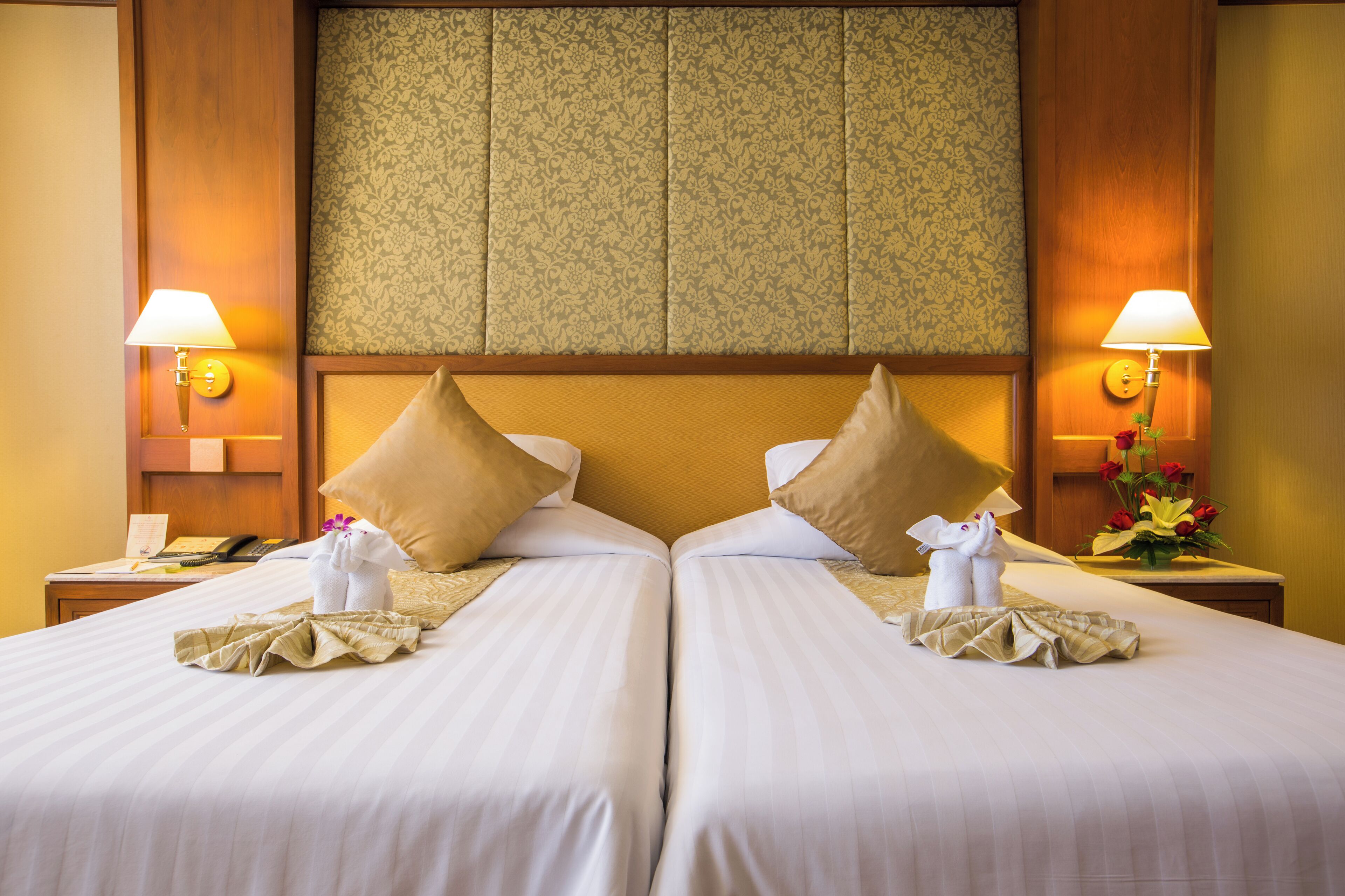 Habitación de hotel con dos camas, cada una con una almohada, una mesita de noche, una lámpara y un jarrón con flores.