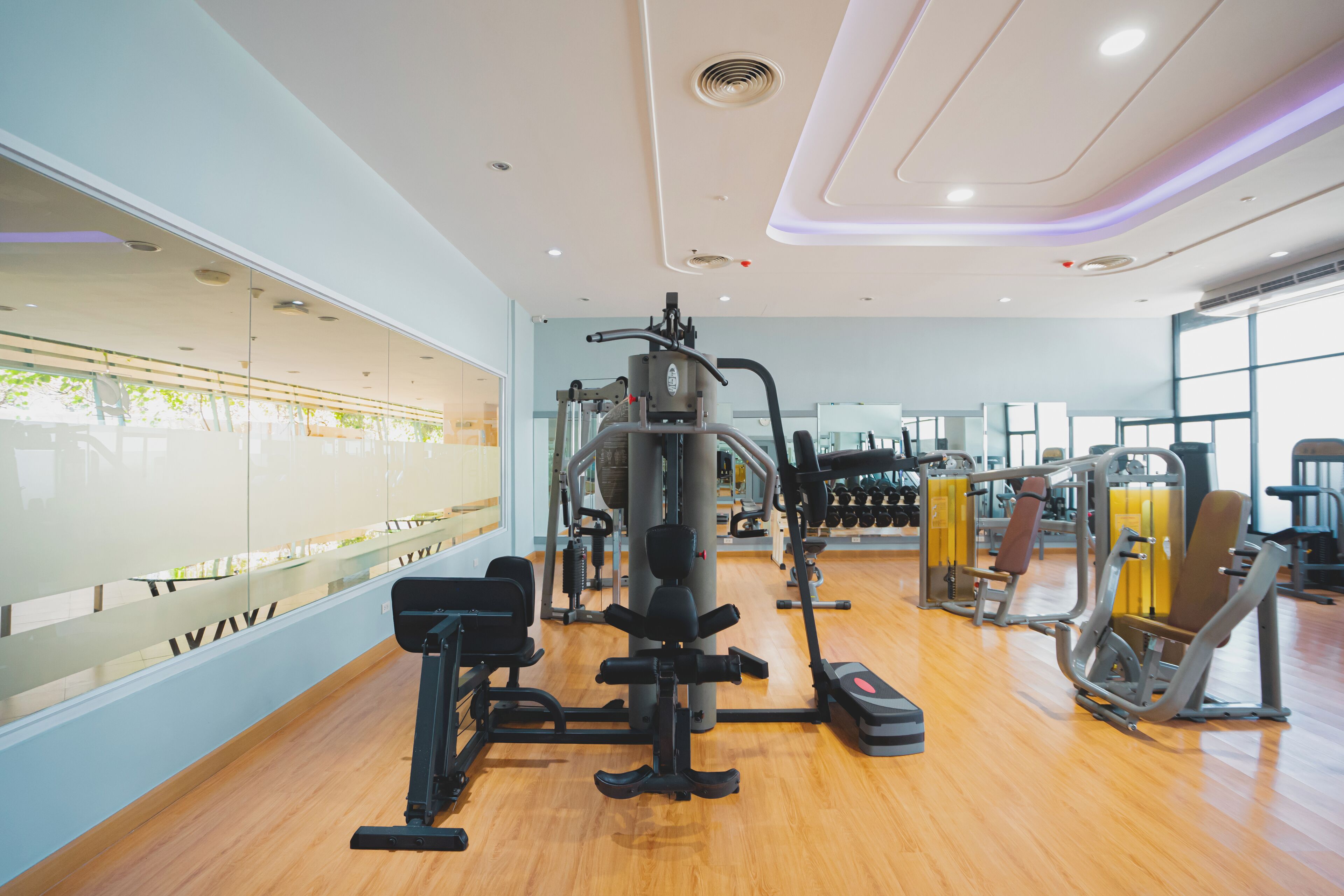A modern gym with various exercise equipment, large windows, and a wooden floor.