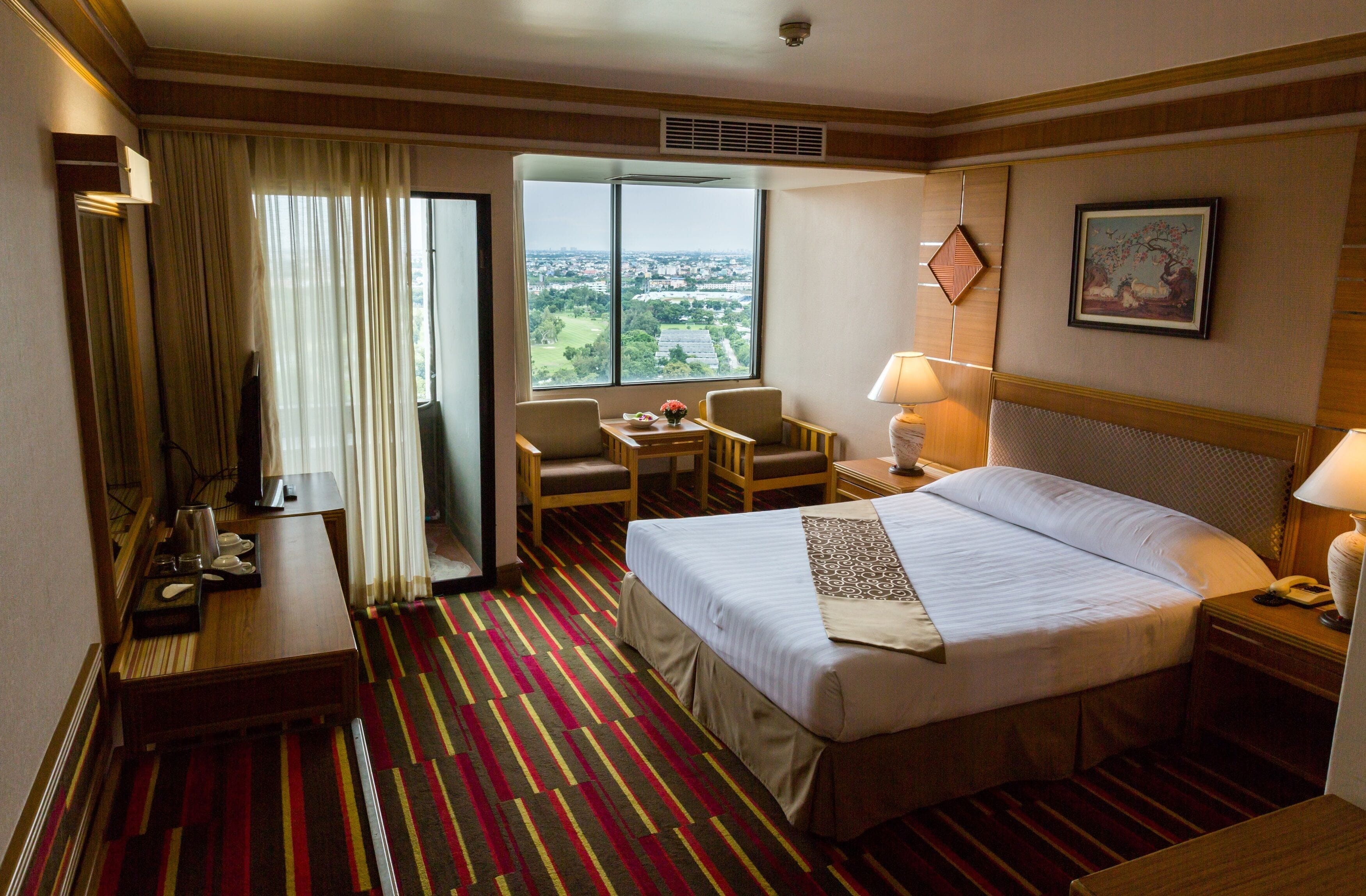 A hotel room with a large bed, a desk, a chair, a small table, a lamp, a telephone, a window with a view of the city, and a painting on the wall.