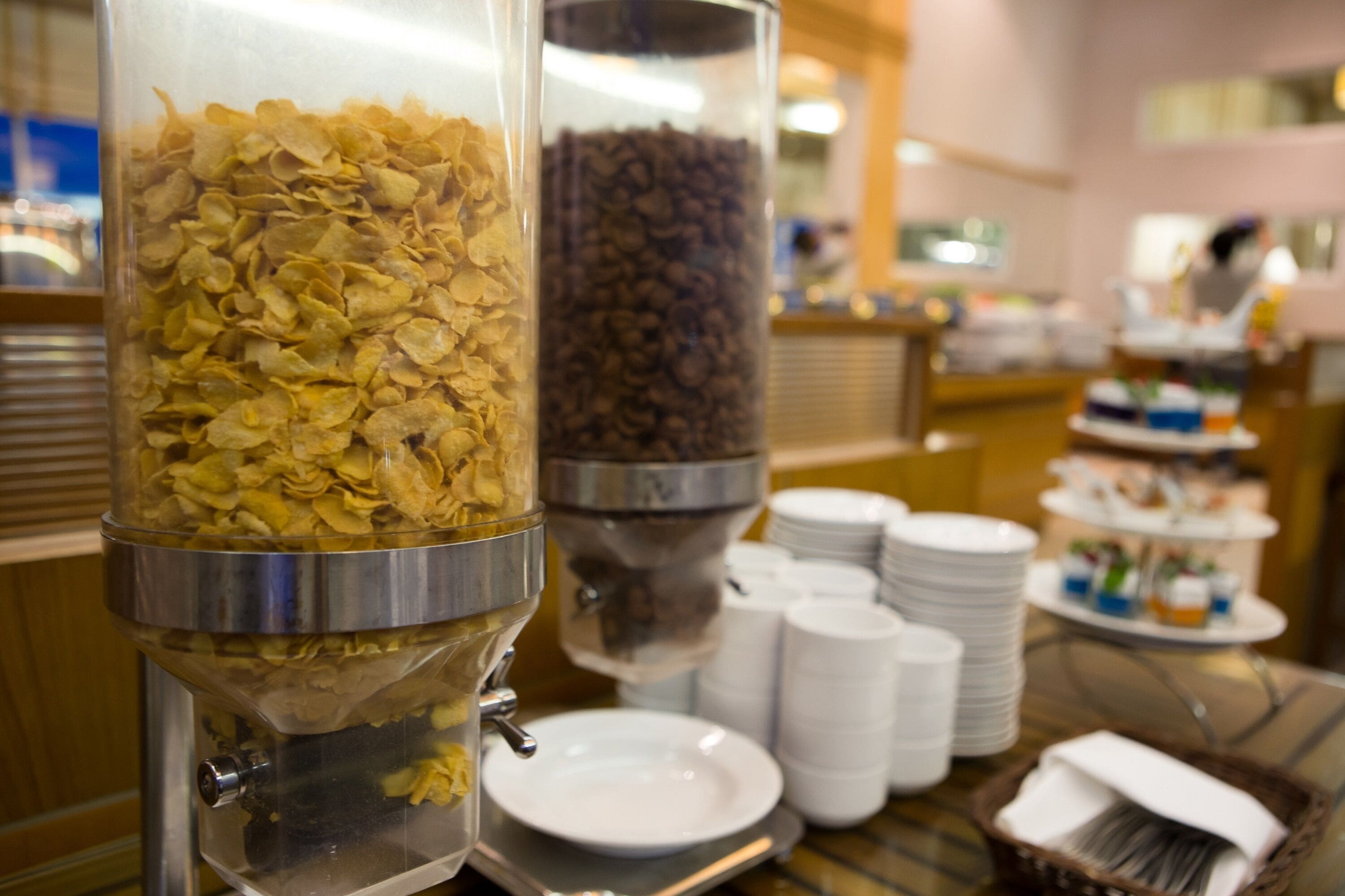 A buffet area with two dispensers, one for cereal flakes and one for coffee beans, and a stack of white plates and cups.