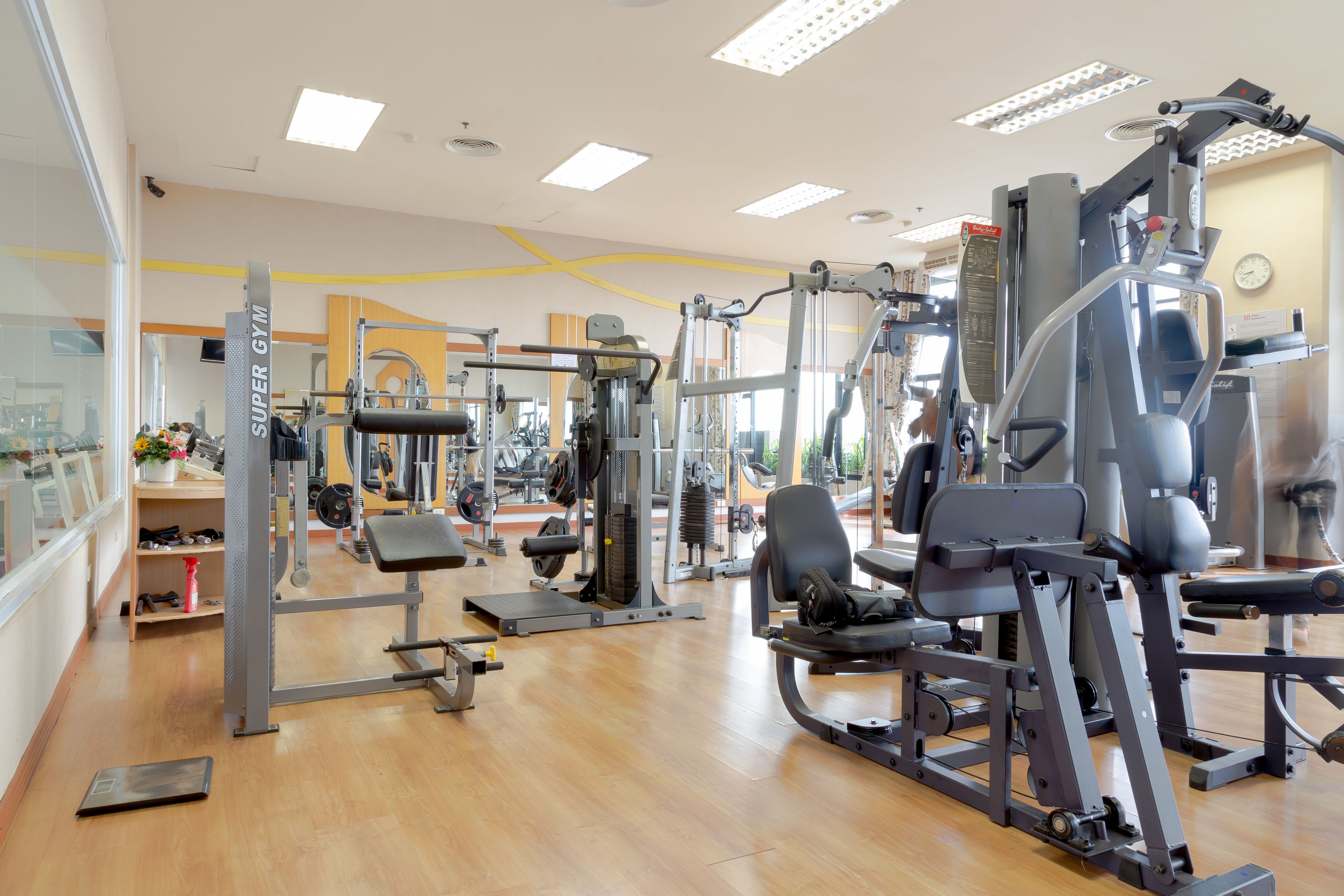 A gym with various exercise machines, wooden flooring, and large windows allowing natural light.