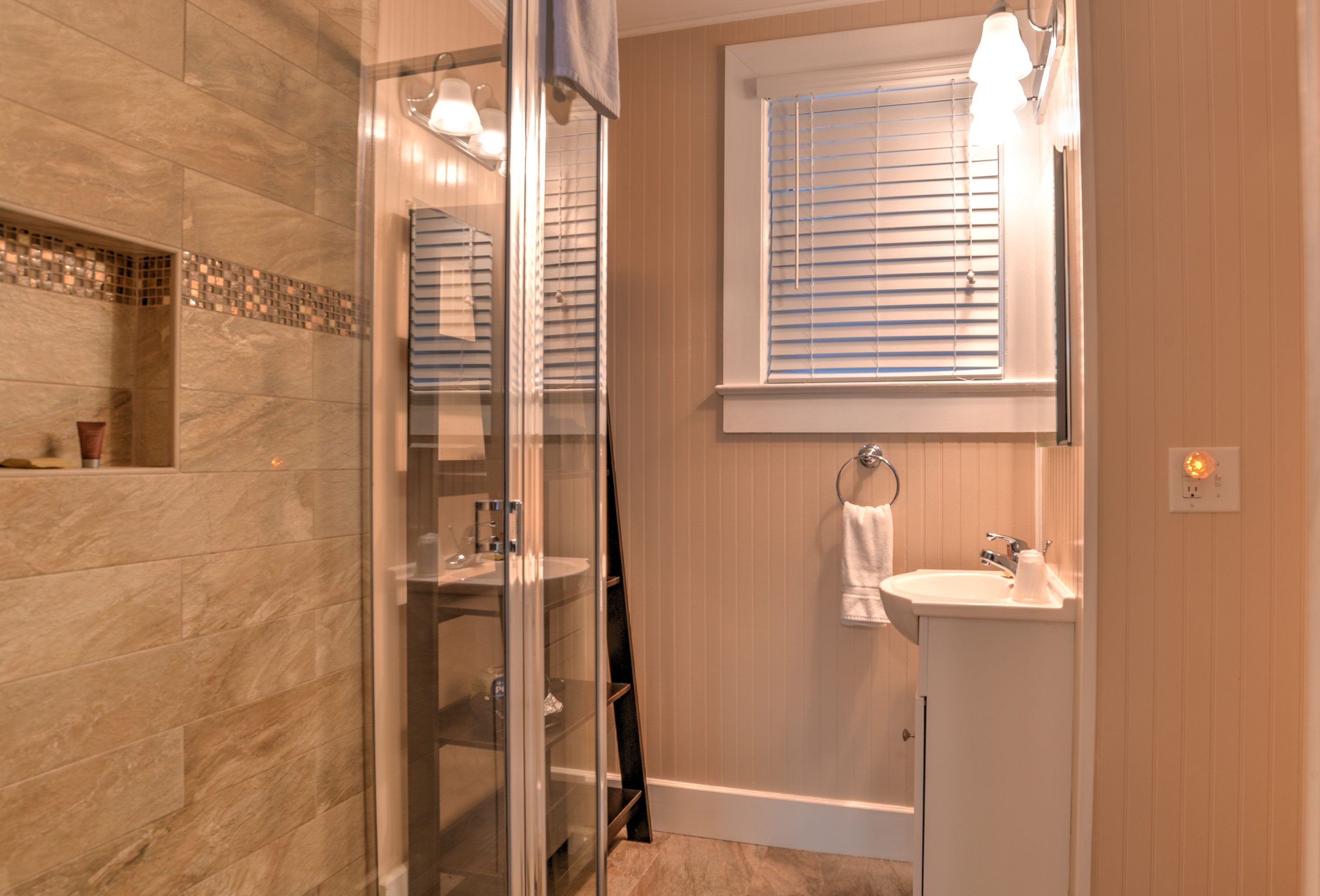 Un baño moderno con cabina de ducha de vidrio, tocador con lavabo y ventana con persianas.