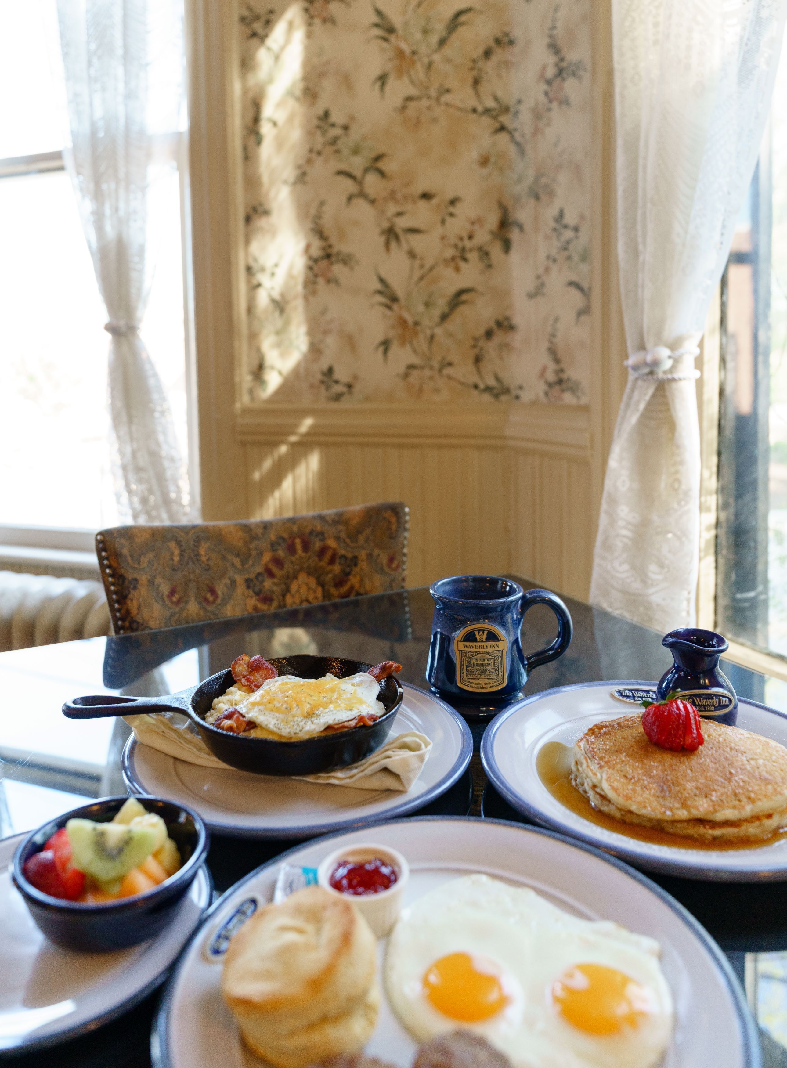 A breakfast table with scrambled eggs, bacon, pancakes, and a muffin, along with a coffee mug and a fruit bowl.