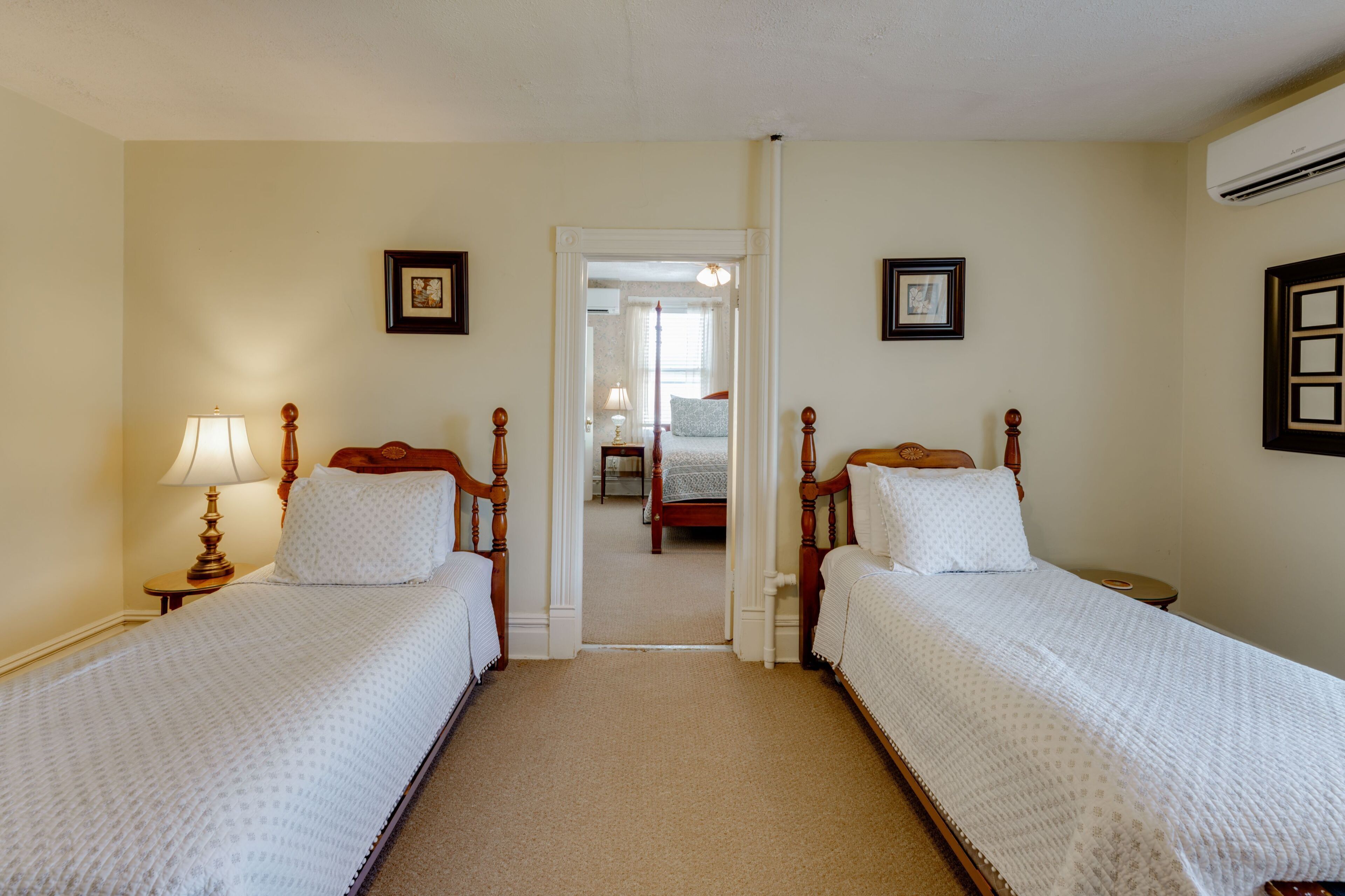 A hotel room with two single beds, a nightstand with a lamp, a framed picture on the wall, and an air conditioning unit.