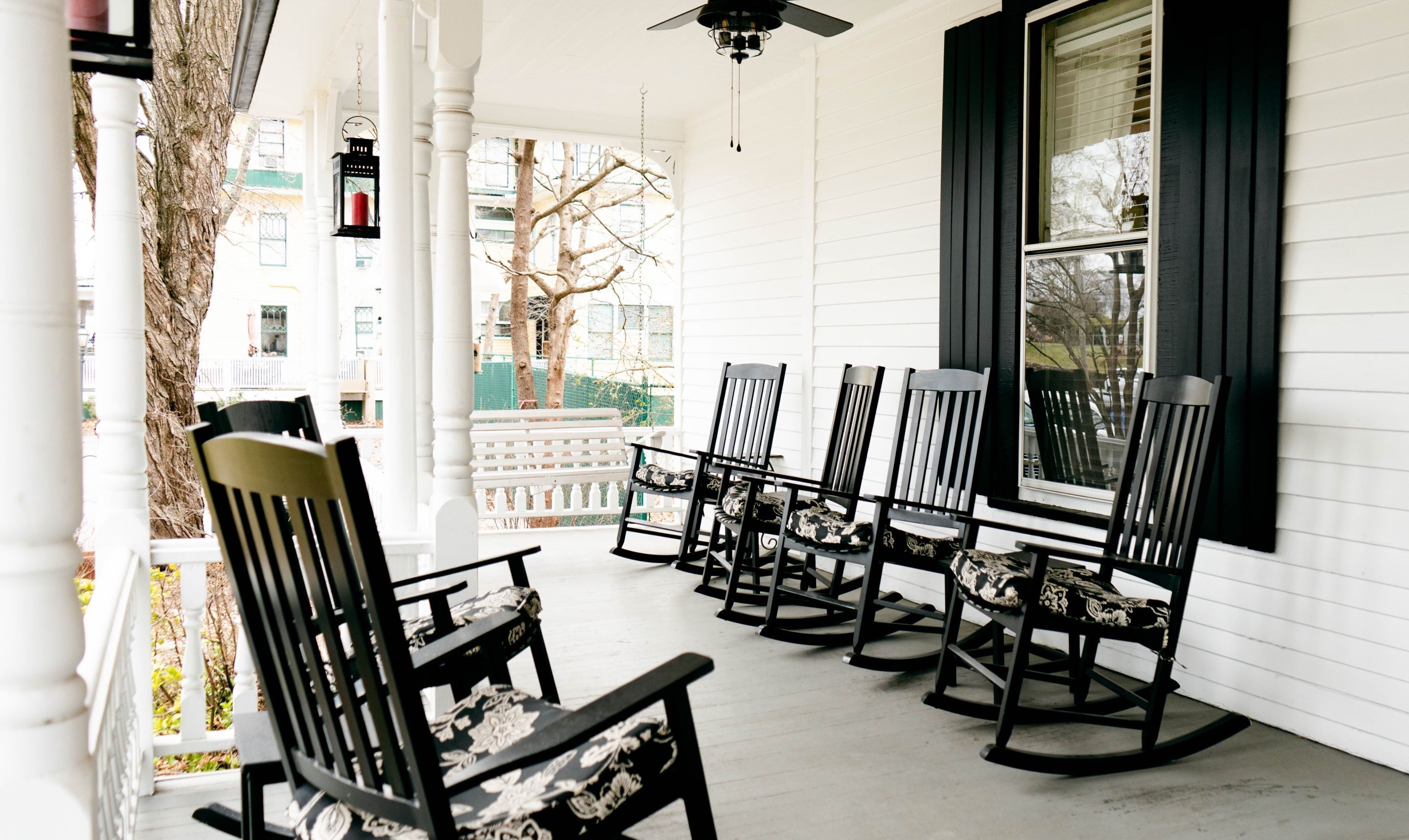 A porch with six rocking chairs, white columns, and a view of a tree and buildings.