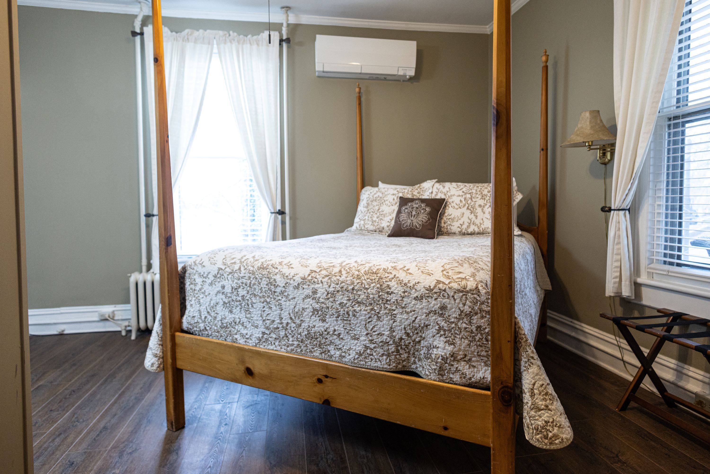 A four-poster bed with a floral bedspread, a wooden floor, a radiator, and a window with blinds.