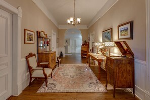 A hallway with a wooden floor, a patterned rug, a chandelier, and a desk with a lamp. - 1898 Waverly Inn (Hendersonville)