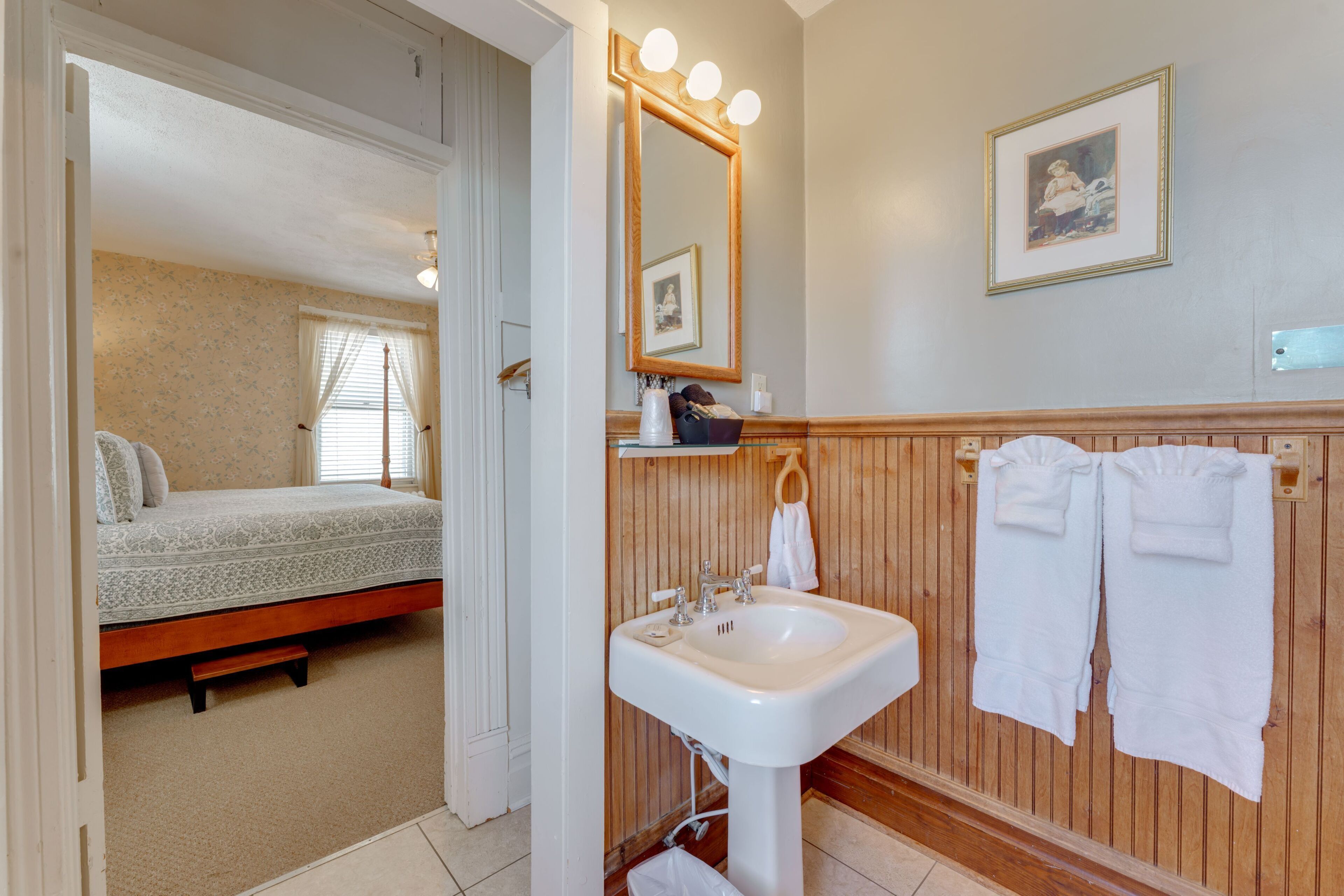 A bedroom with a bed, a bathroom with a sink and mirror, wooden paneling, and a framed picture on the wall.