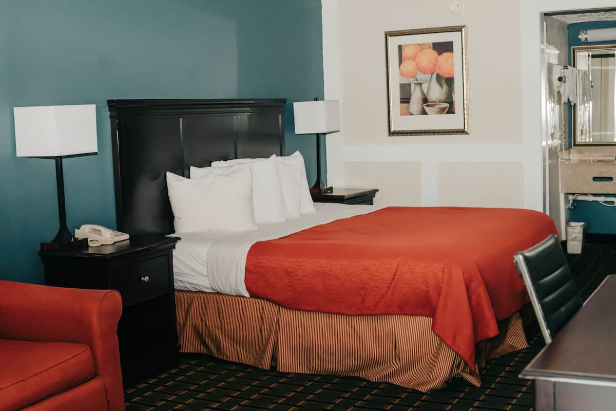 A hotel room with a bed, a nightstand, a telephone, a chair, and a bathroom with a mirror and towel rack.