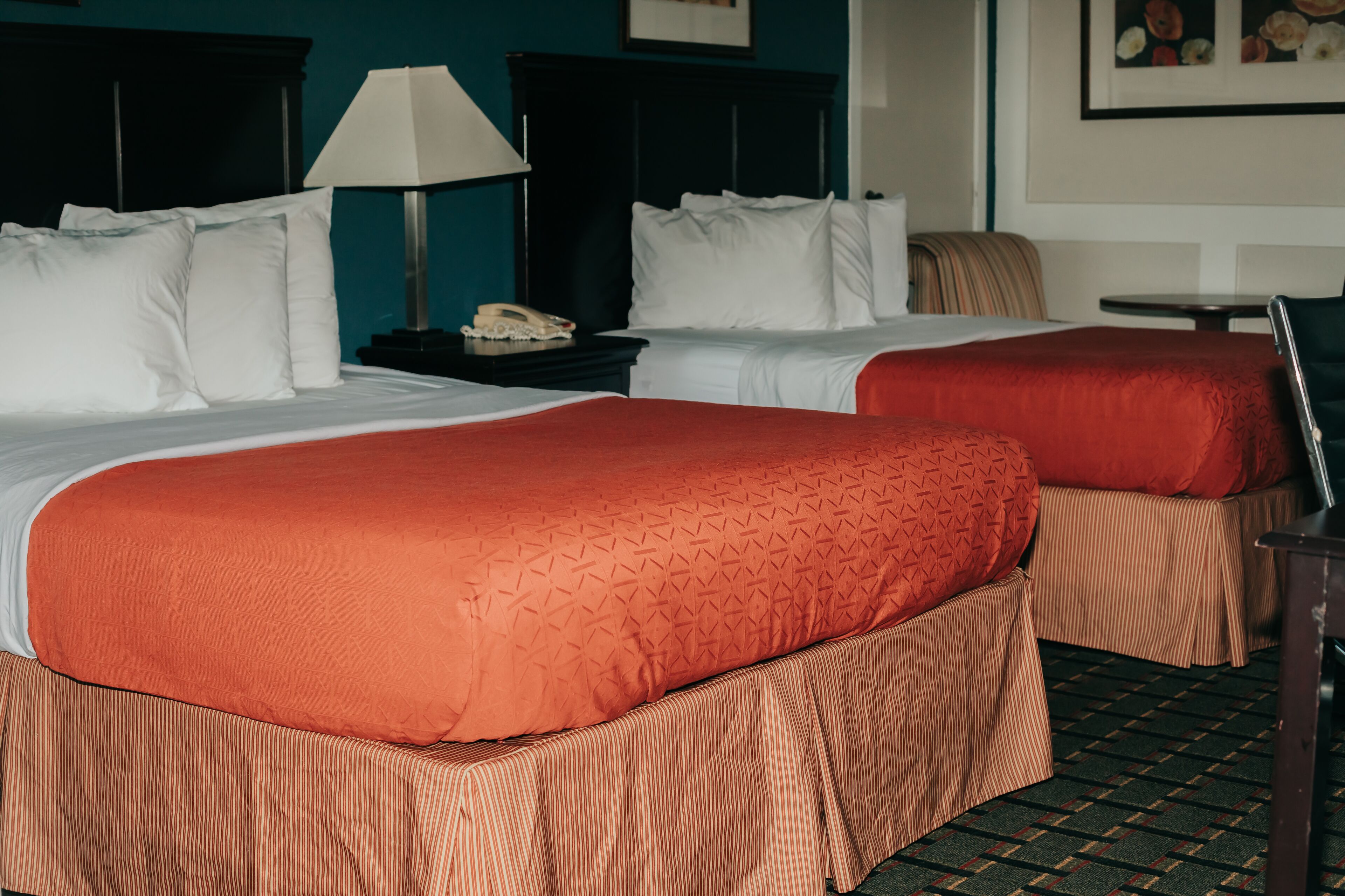 Habitación de hotel con dos camas, cada una con ropa de cama blanca y naranja, una mesita de noche con una lámpara y un cuadro en la pared.