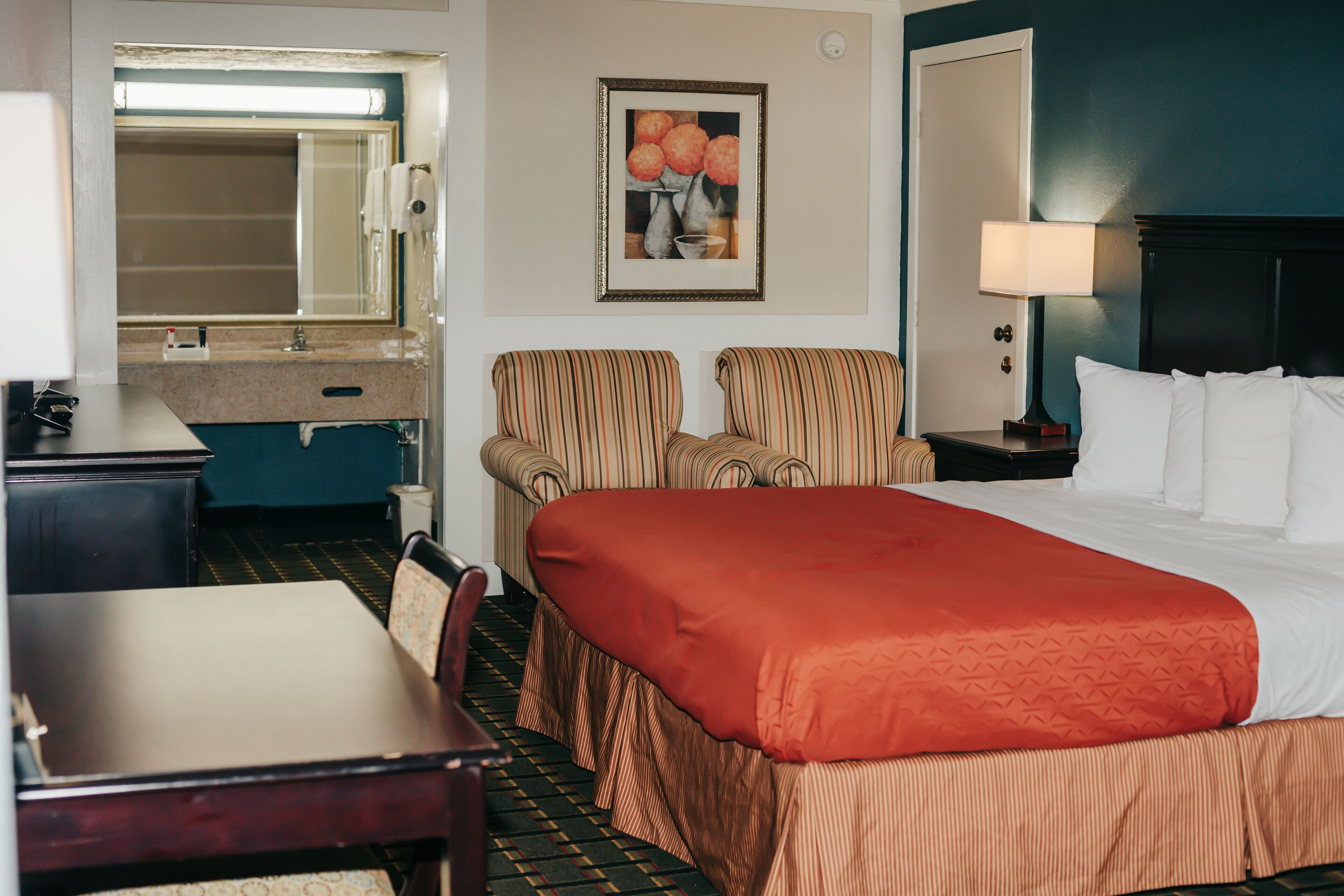 A hotel room with a bed, two striped armchairs, a desk, and a nightstand with a lamp.