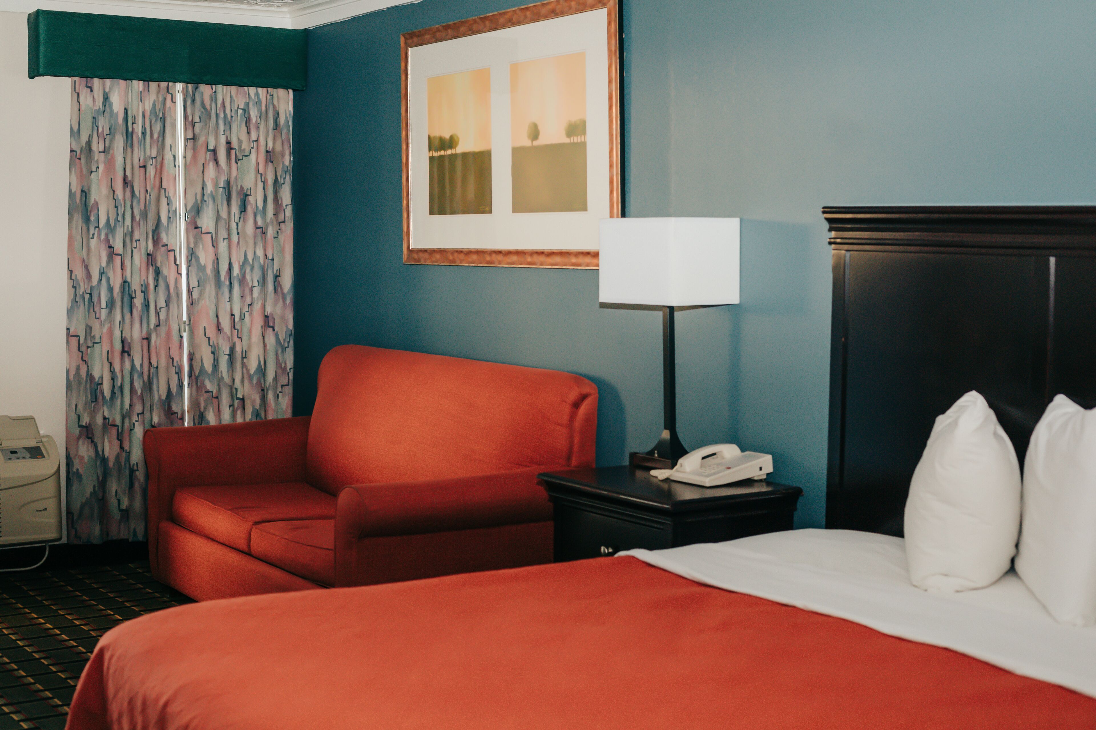 A hotel room with a bed, a red armchair, a nightstand with a lamp, and a telephone.