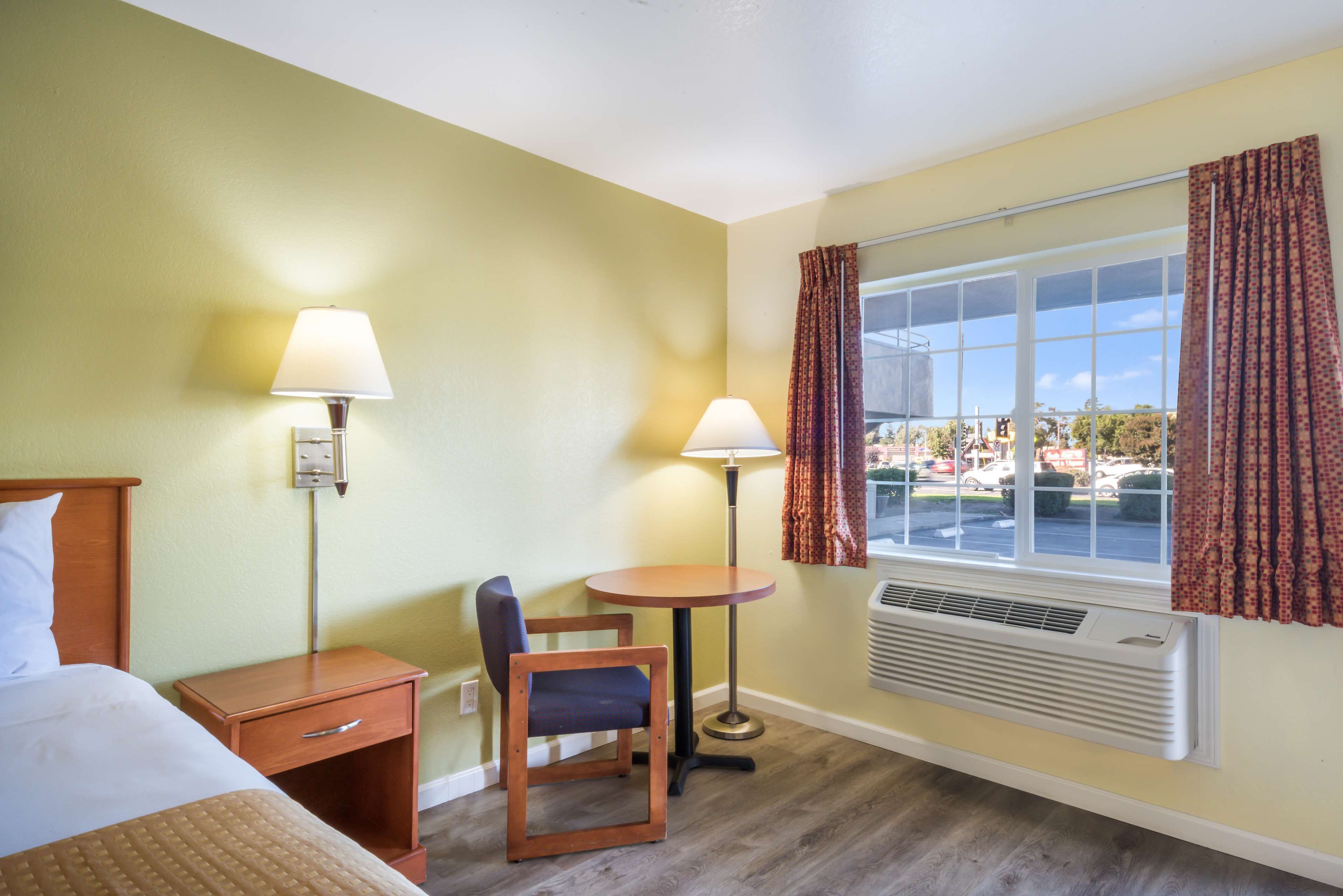 Habitación de hotel con cama, escritorio, silla, dos lámparas, ventana con cortinas y un equipo de aire acondicionado.