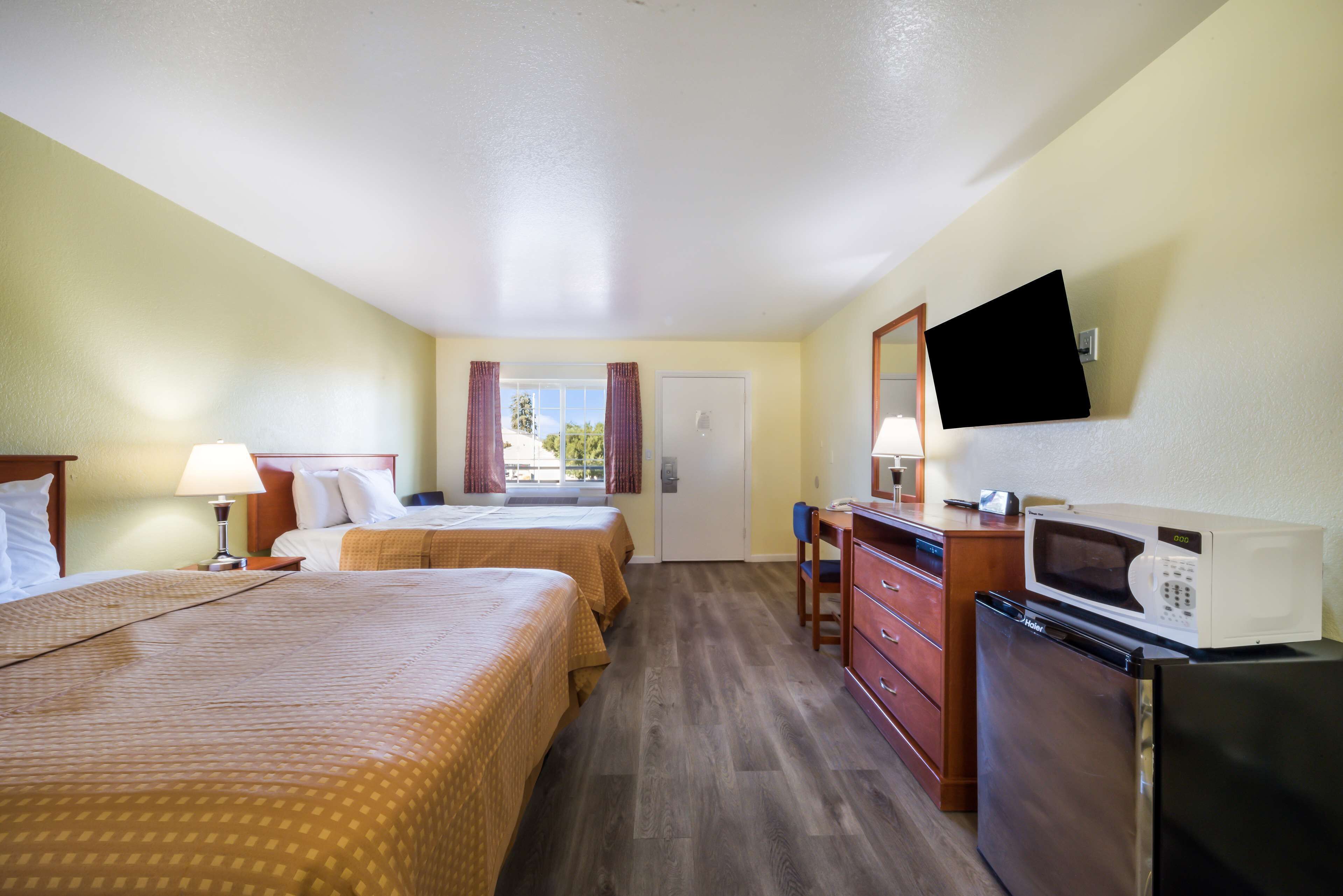 A hotel room with two beds, a desk, a chair, a dresser, a microwave, a television, and a window with curtains.