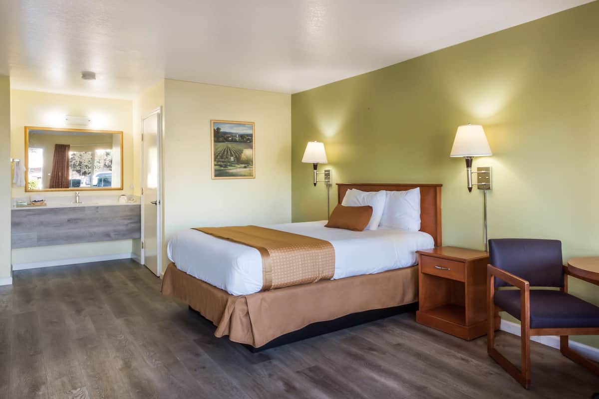 A hotel room with a bed, bedside table, chair, and a bathroom with a mirror and sink.