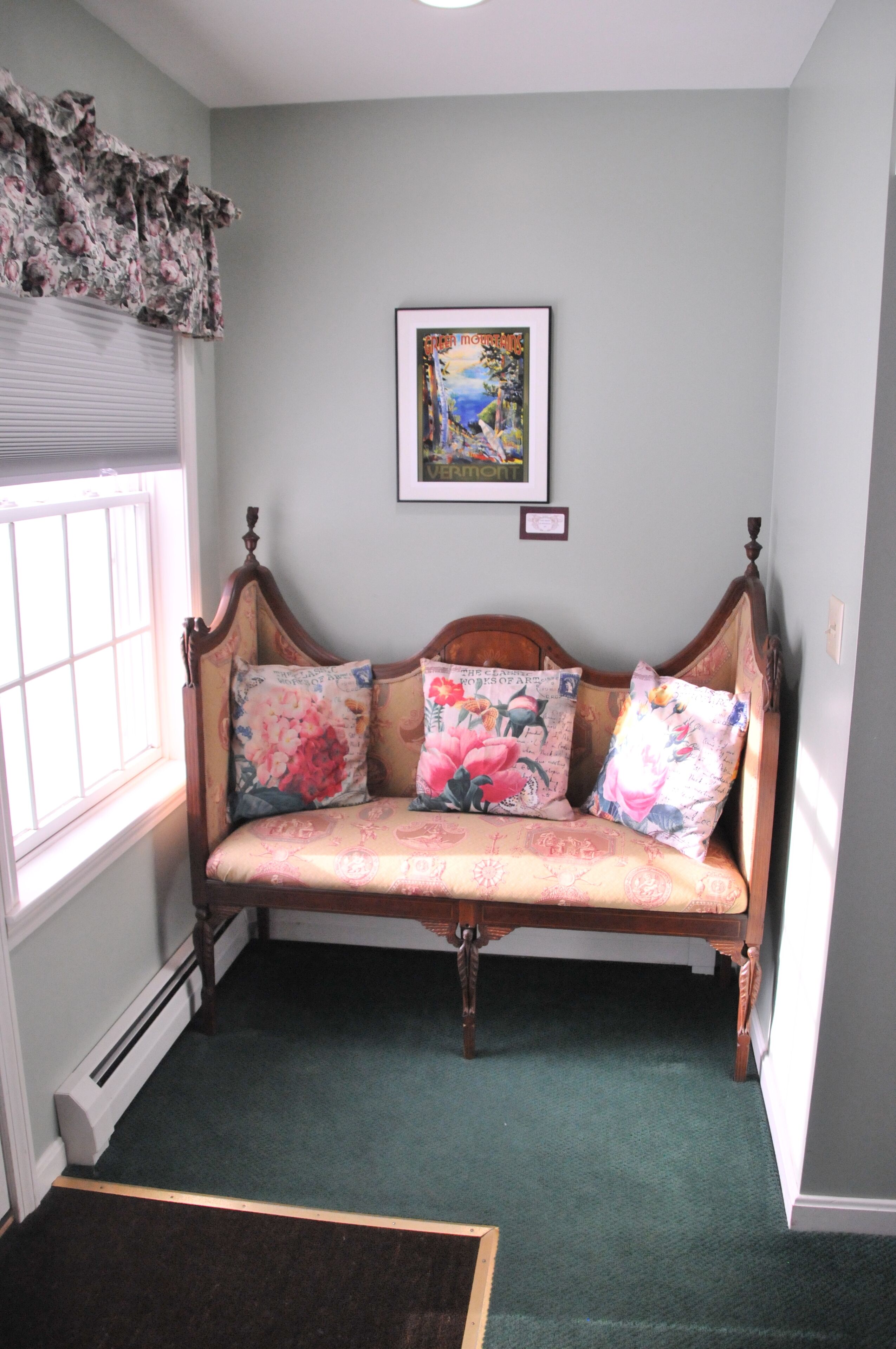 A room with a wooden bench, floral cushions, a framed artwork on the wall, and a window with blinds.