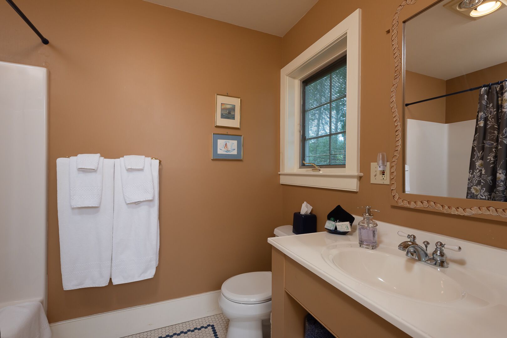 Un baño con pared color beige, un lavamanos blanco, un inodoro y una ventana con persianas. Hay dos cuadros enmarcados en la pared y un perchero con dos toallas.