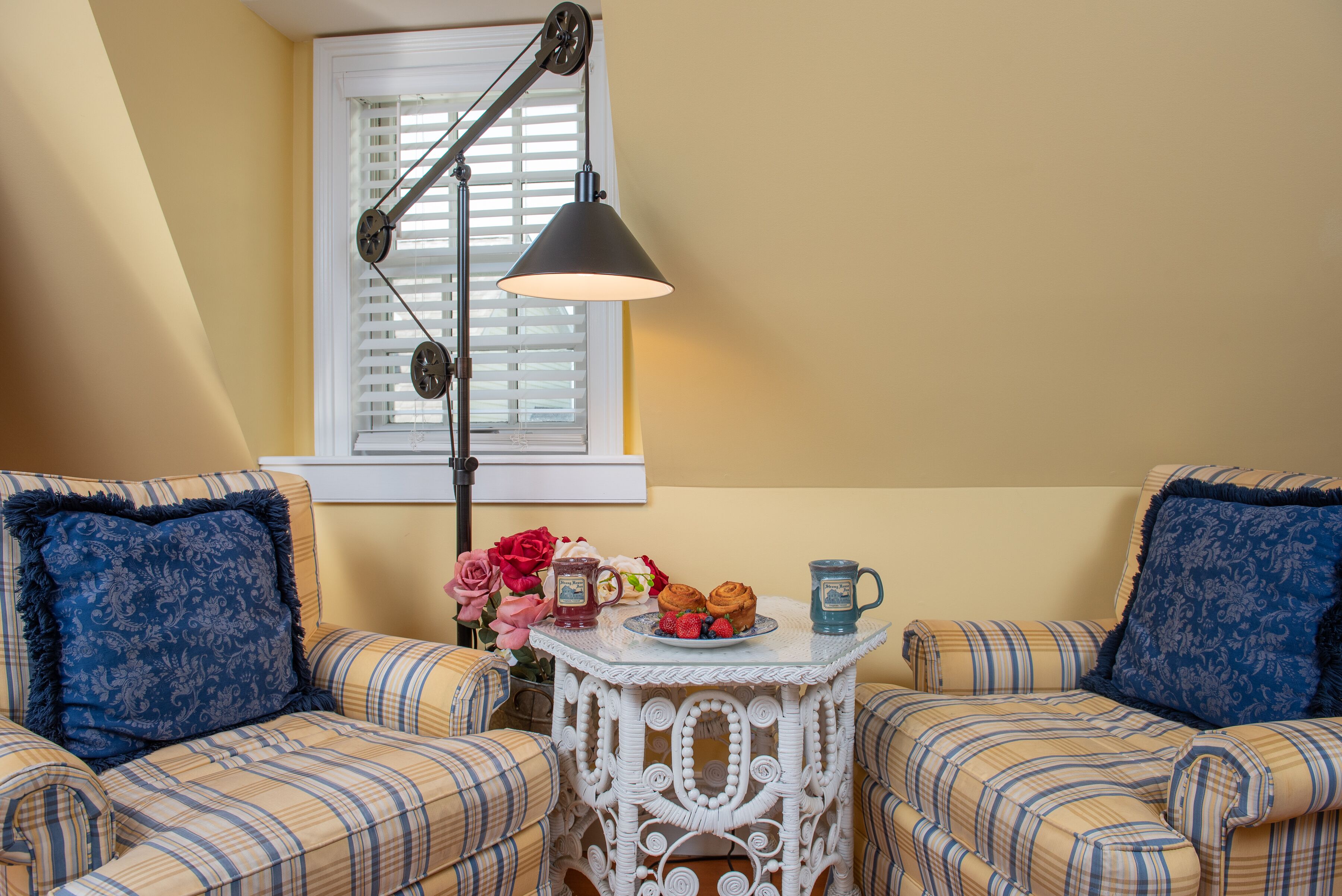 A cozy room with two plaid sofas, a small white table with a plate of food and drinks, and a floor lamp.