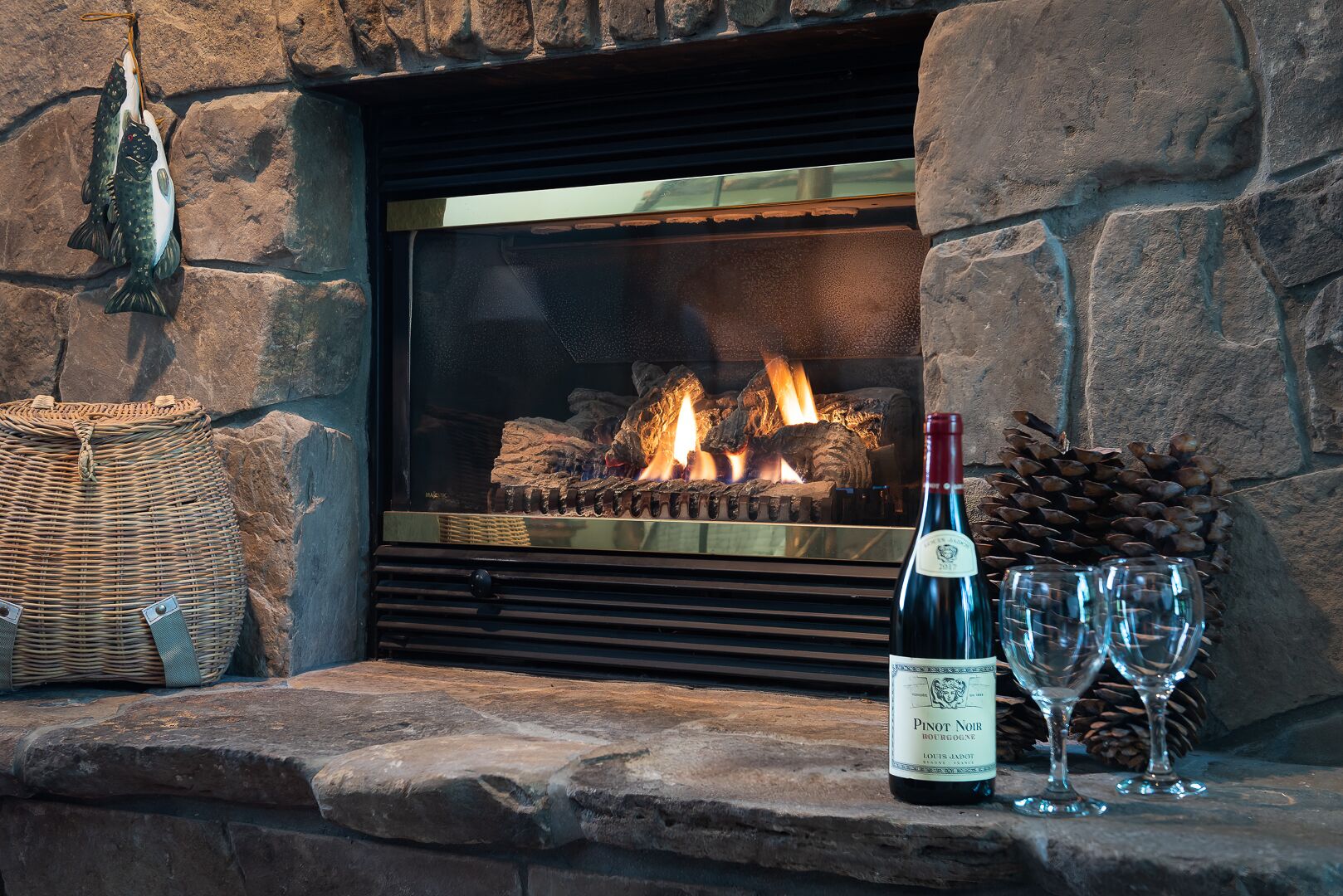 A stone fireplace with a log fire, a bottle of Pinot Noir, and two glasses on a stone ledge.