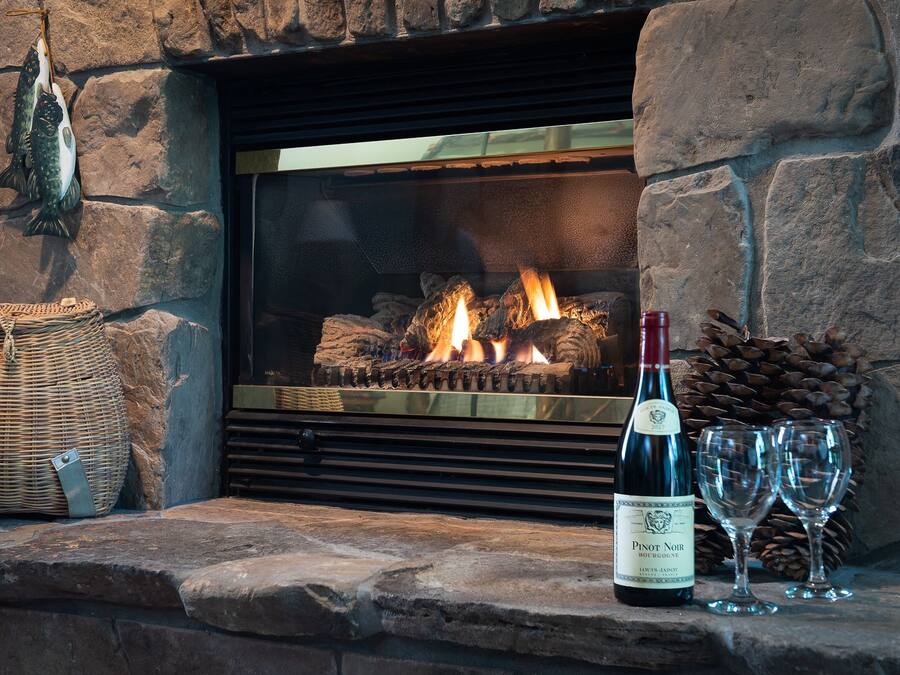 A stone fireplace with a log fire, a bottle of Pinot Noir, and two glasses on a stone ledge.