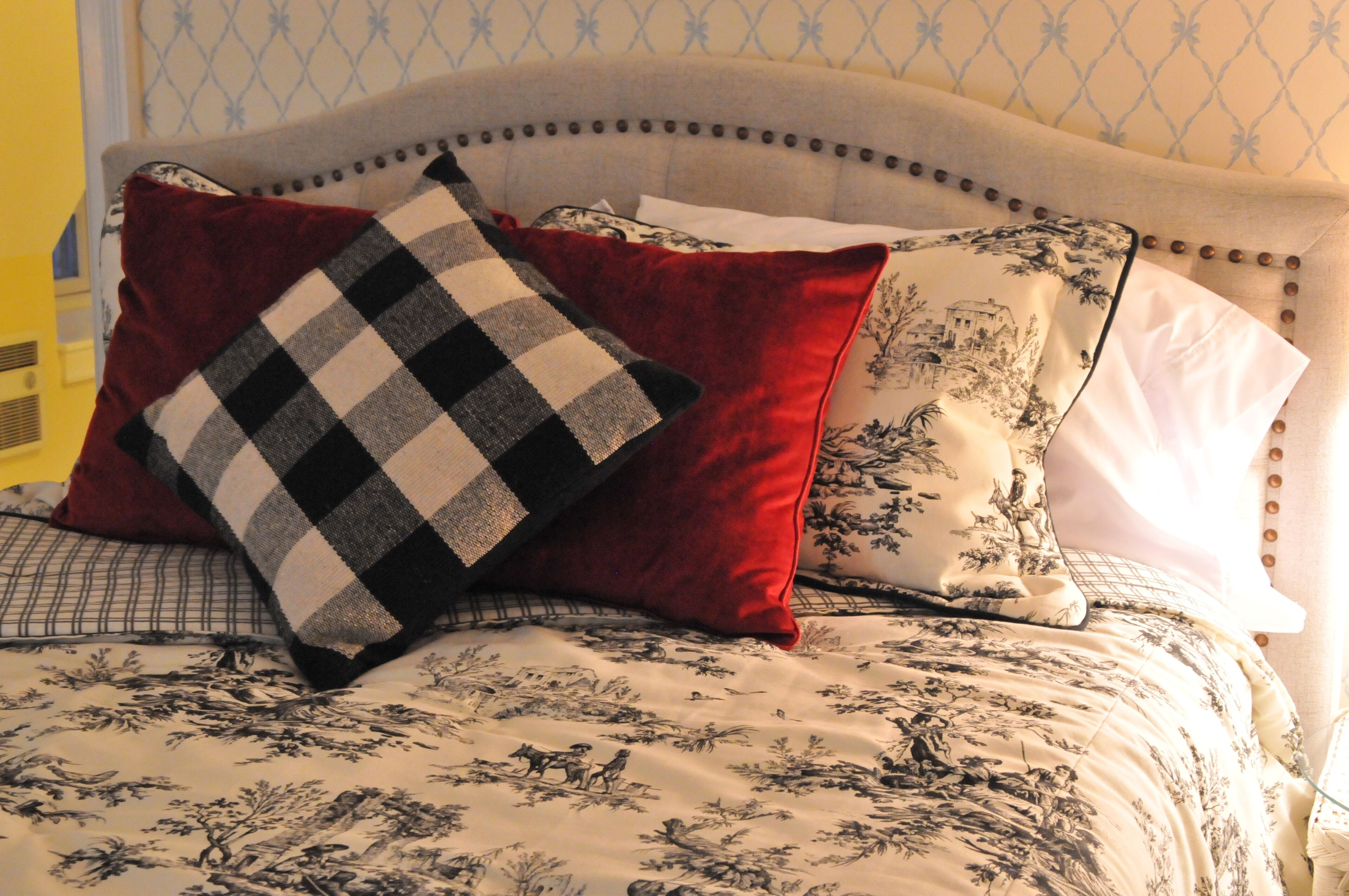 A bed with a floral patterned bedspread, a checkered pillow, and a red pillow.