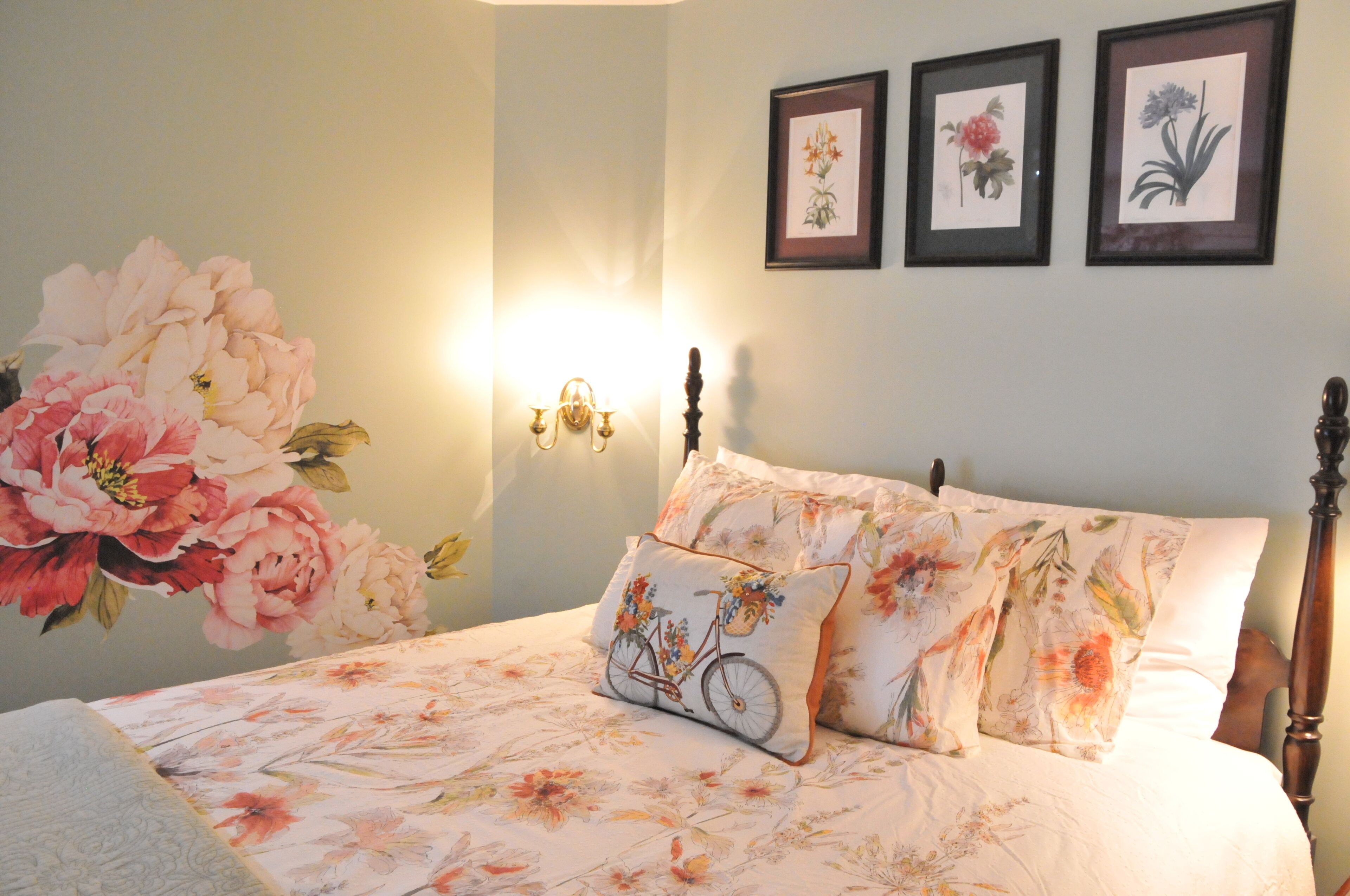 A bedroom with a floral bedspread, a bicycle-themed pillow, and three framed floral pictures on the wall.