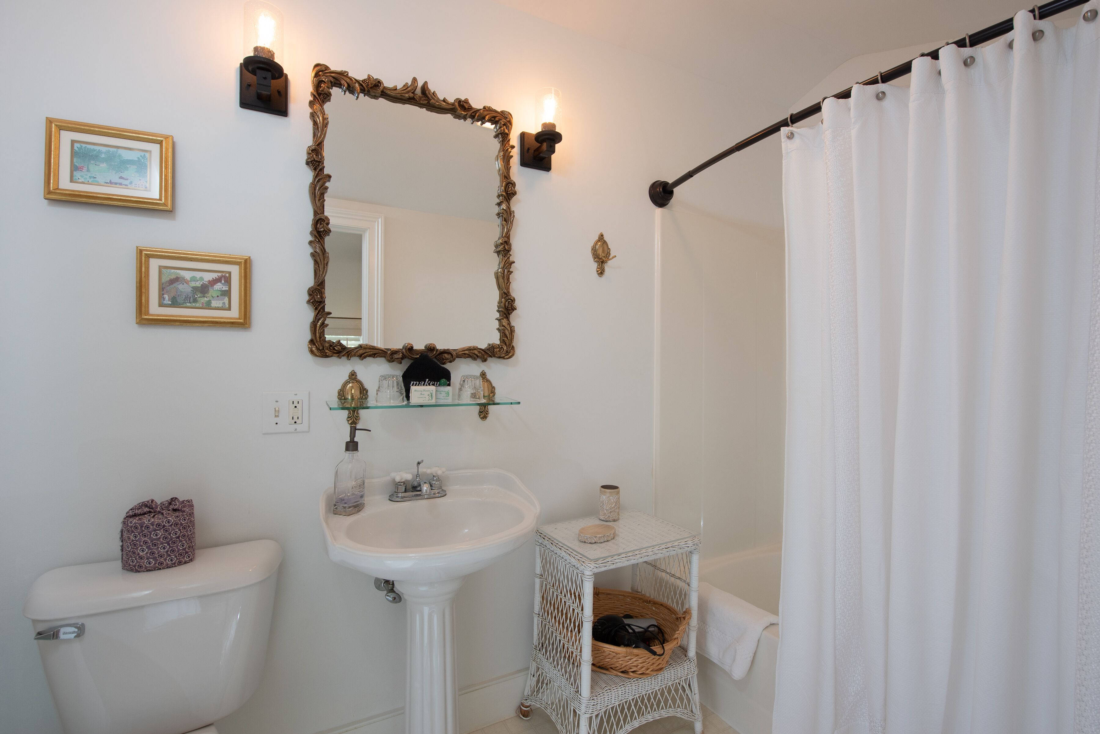 A bathroom with a white toilet, pedestal sink, and a small wicker basket on a white wicker shelf.