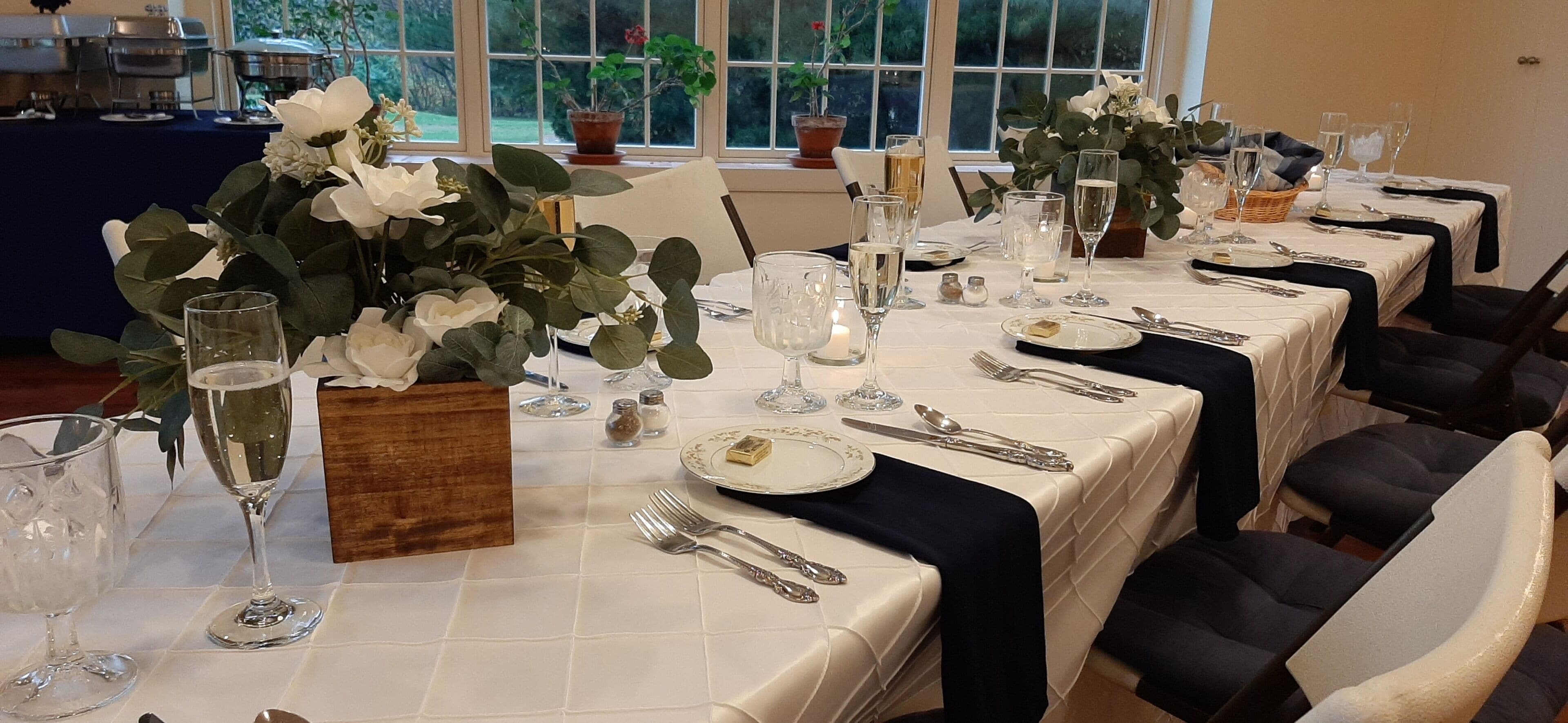 A dining table set with white tablecloths, silverware, and glassware, with potted plants and a candle centerpiece.