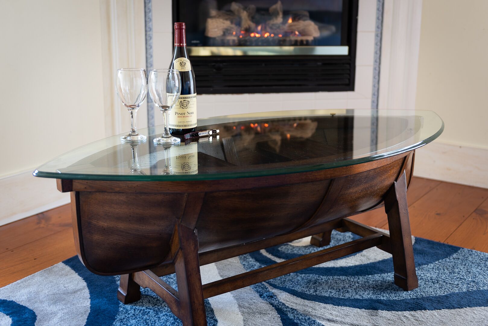 A glass-top wooden table with a bottle of wine and two glasses on it, placed in a room with a fireplace in the background.