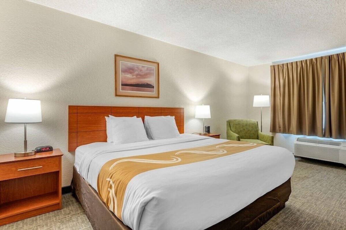 A hotel room with a bed, two bedside tables, a green armchair, a window with curtains, and a framed picture on the wall.