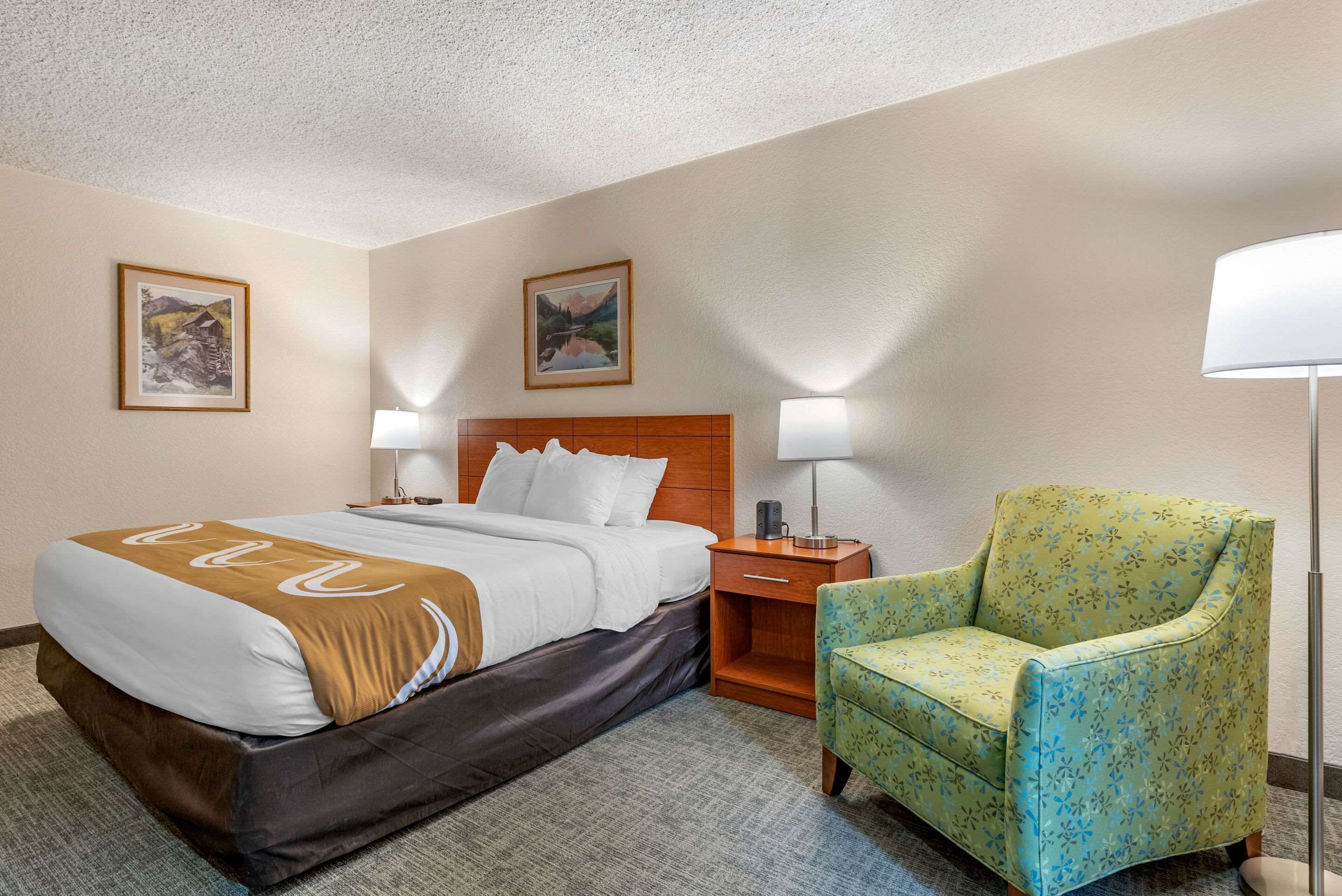 Una habitación de hotel con cama, una silla, una mesita de noche y dos lámparas de pared.
