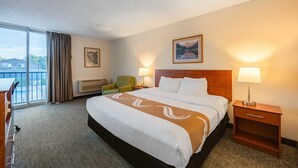 A hotel room with a large bed, a wooden headboard, two bedside lamps, a green armchair, a desk with a chair, and a large window with curtains. - Quality Inn & Suites Canon City (Canon City)