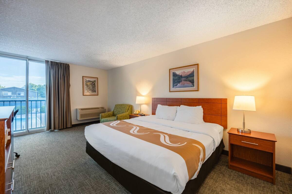A hotel room with a large bed, a wooden headboard, two bedside lamps, a green armchair, a desk with a chair, and a large window with curtains.