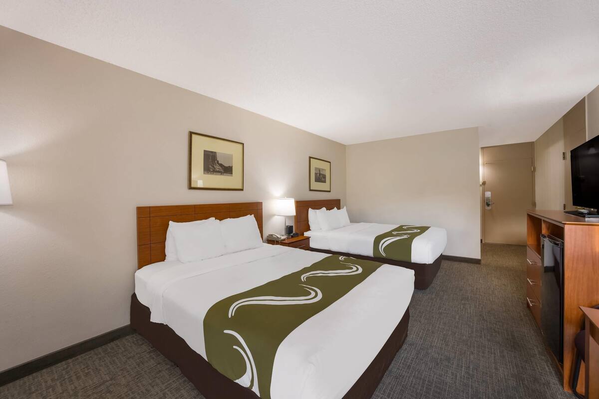 A hotel room with two beds, a desk, a television, and framed pictures on the wall.