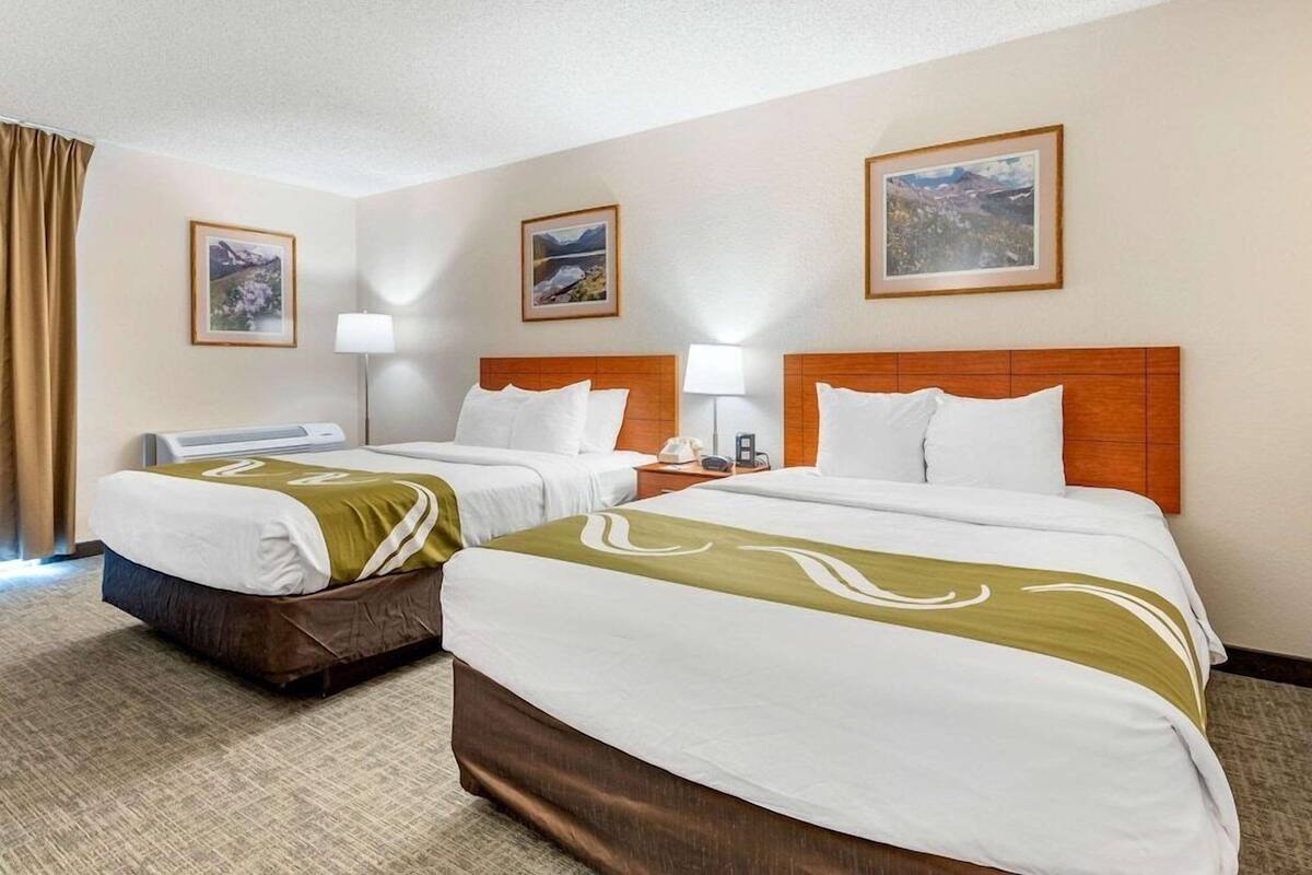 A hotel room with two beds, a desk, a chair, a lamp, and framed pictures on the wall.