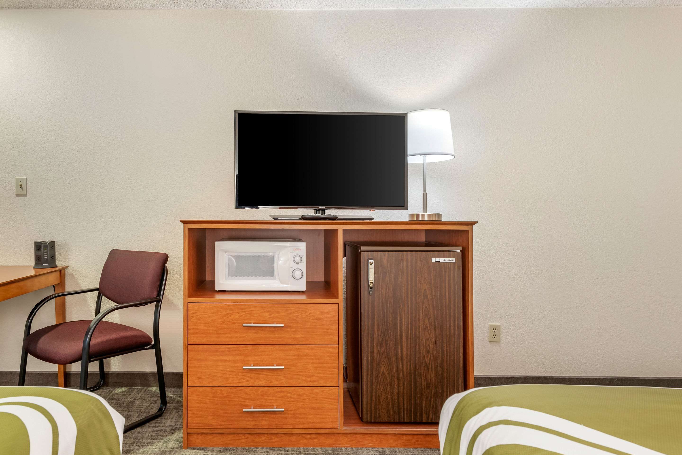 Habitación de hotel con cama, escritorio con silla, televisor, microondas, refrigerador y lámpara.