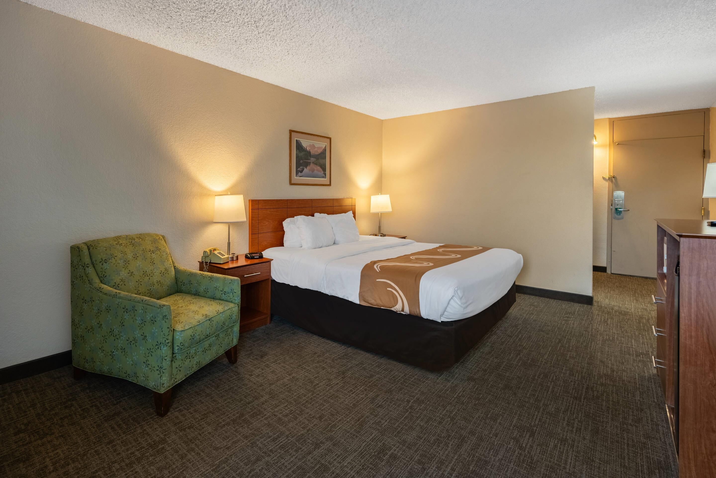 A hotel room with a bed, a green armchair, two bedside lamps, and a wooden nightstand.