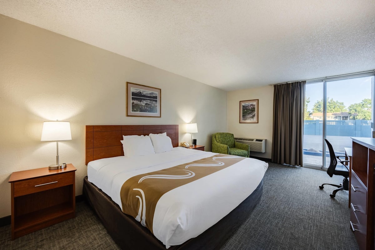 A hotel room with a large bed, two bedside lamps, a green armchair, a desk with a chair, and a large window with a view.