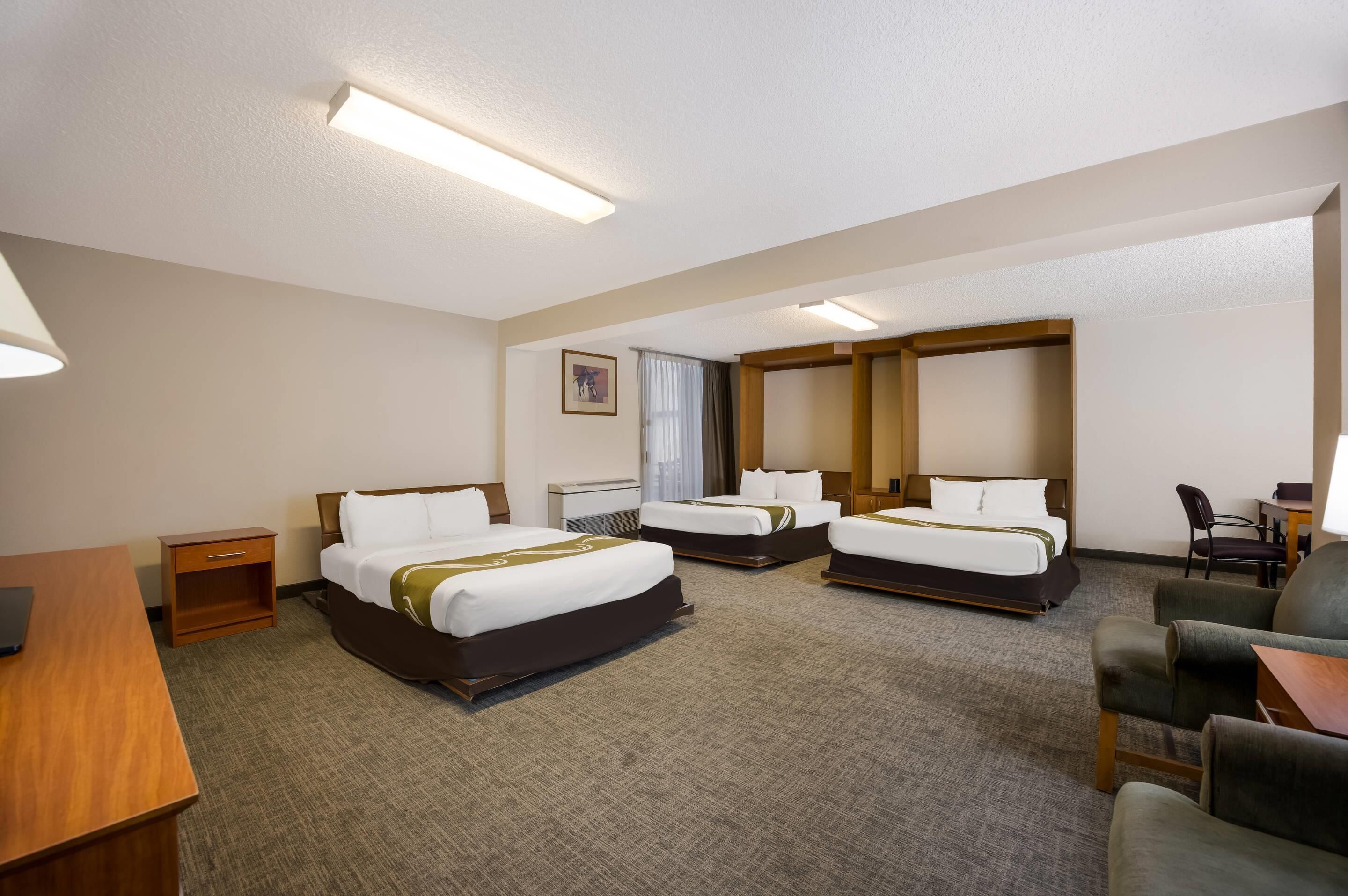 Quarto de hotel com três camas, uma escrivaninha, uma cadeira e uma pequena mesa.