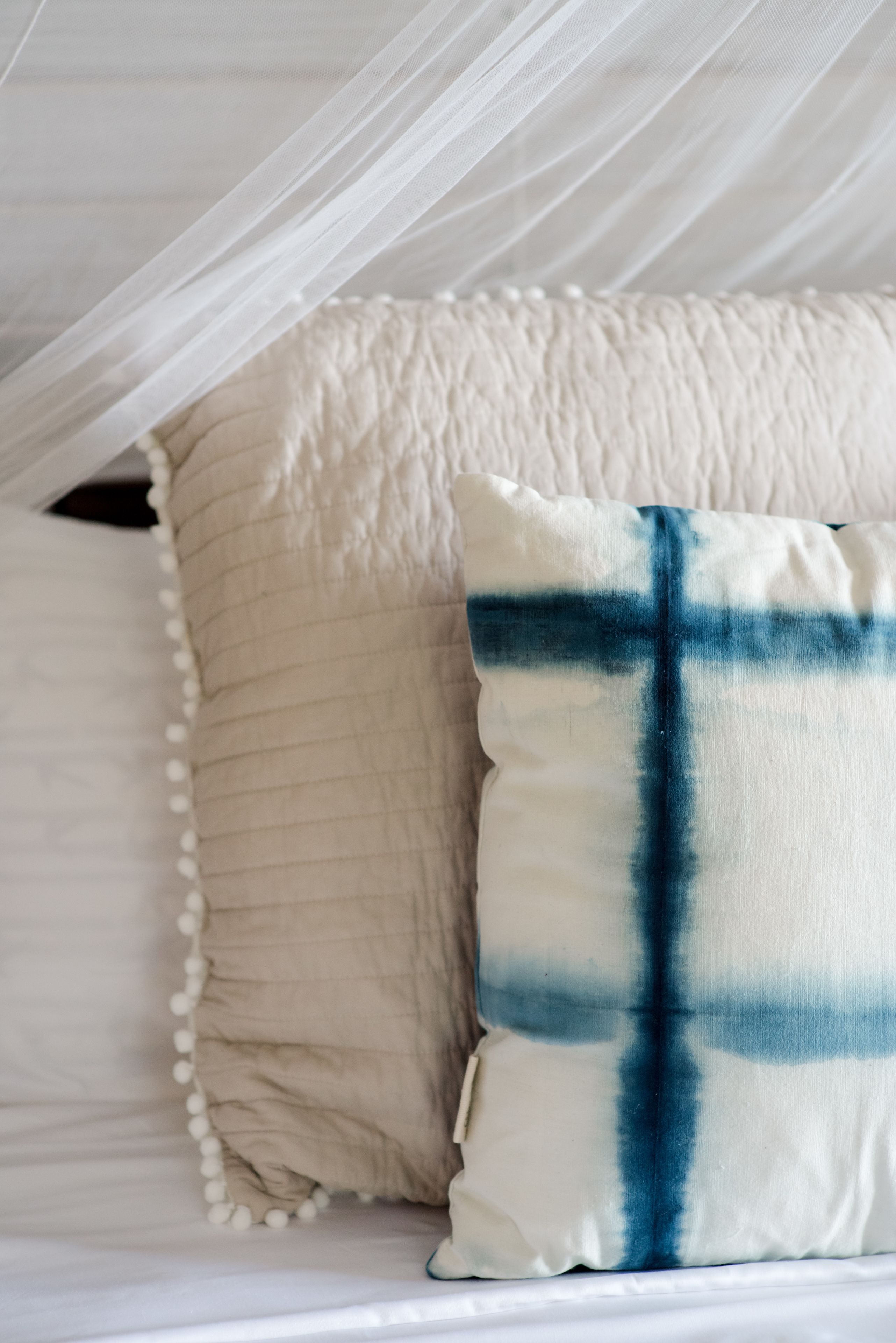 A bed with a white quilted headboard, a beige pillow with a white pom-pom trim, and a blue and white plaid pillow.