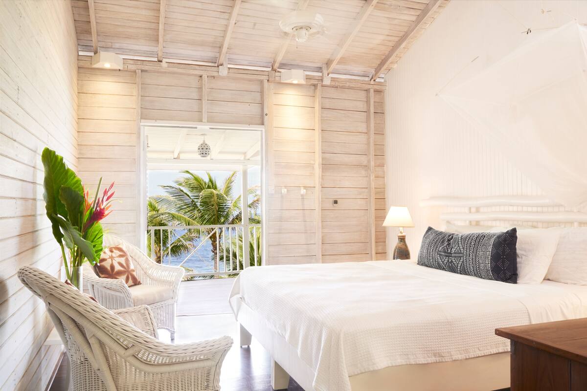 A bedroom with a bed, a wicker chair, a nightstand, and a view of the ocean.