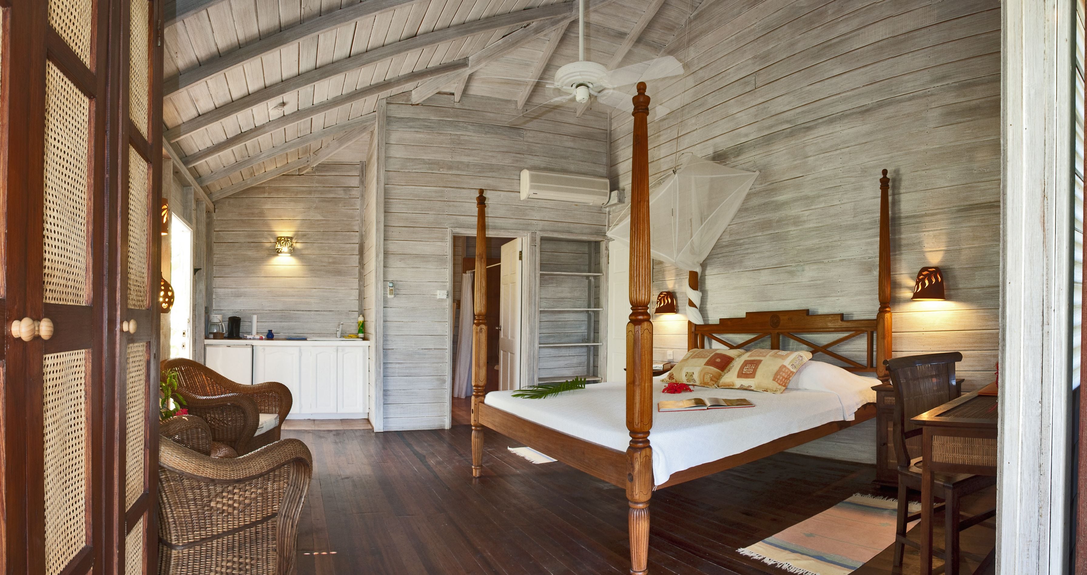 A four-poster bed with a wooden headboard, a dining table with chairs, wicker furniture, and a kitchen area with white cabinets.